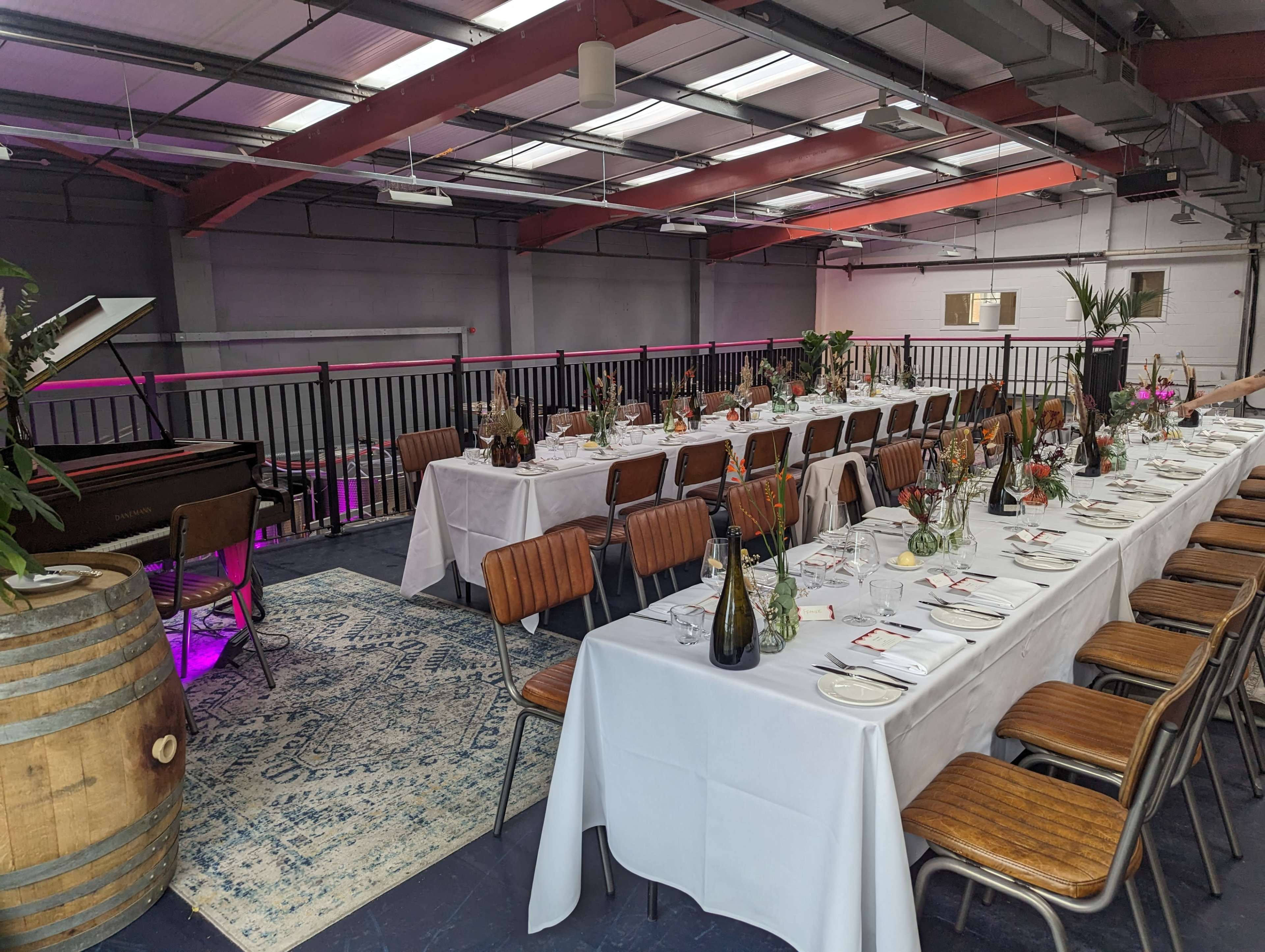 A spacious dining area features a long table set for a formal meal, with green plants and elegant tableware, alongside a piano in a well-lit venue.