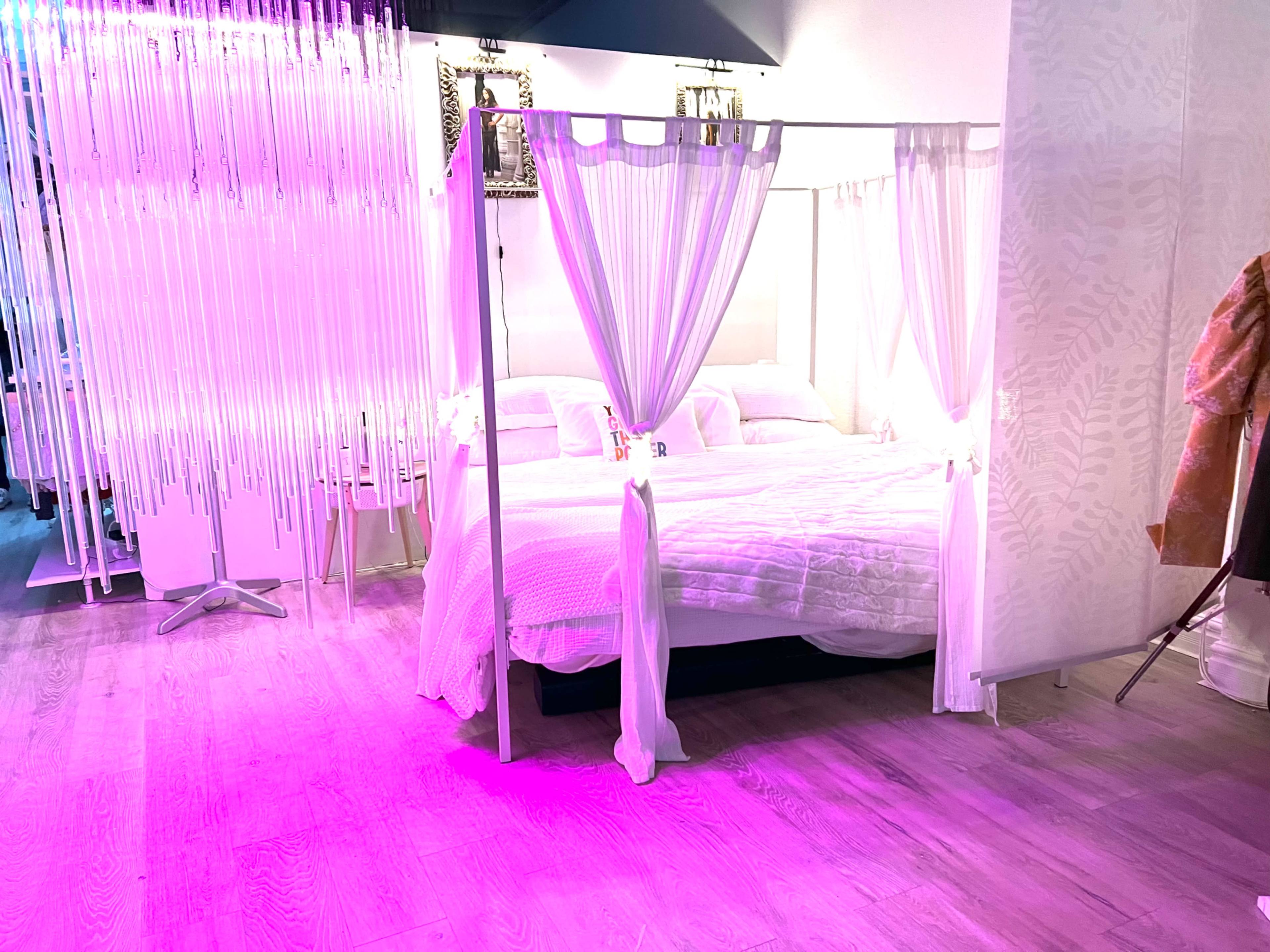 The image shows a bedroom with a four-poster bed draped in white curtains and illuminated by purple lighting.