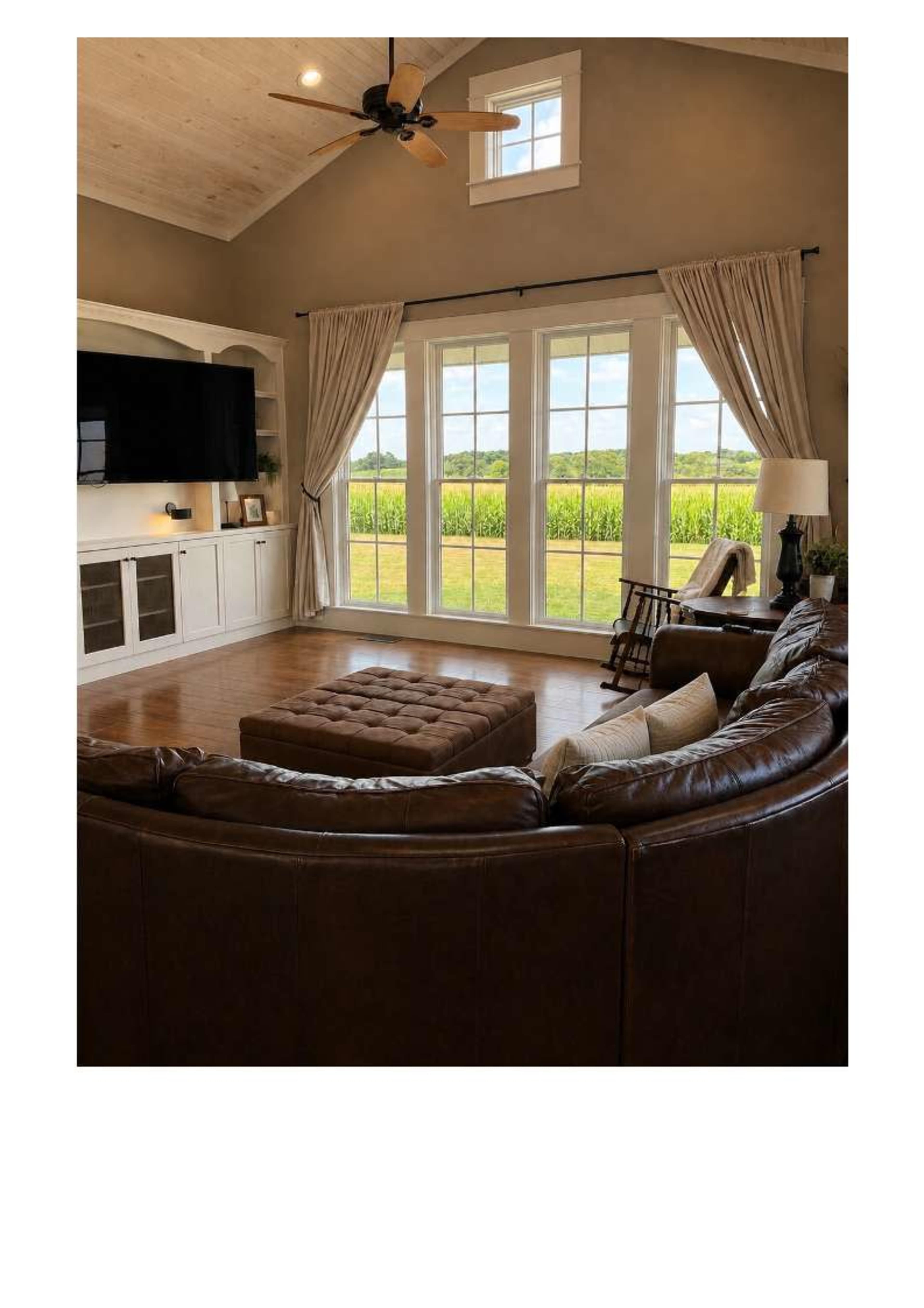 The image shows a cozy living room with a brown leather sectional sofa, a large window displaying a view of a green field, and a decorative coffee table in the center.