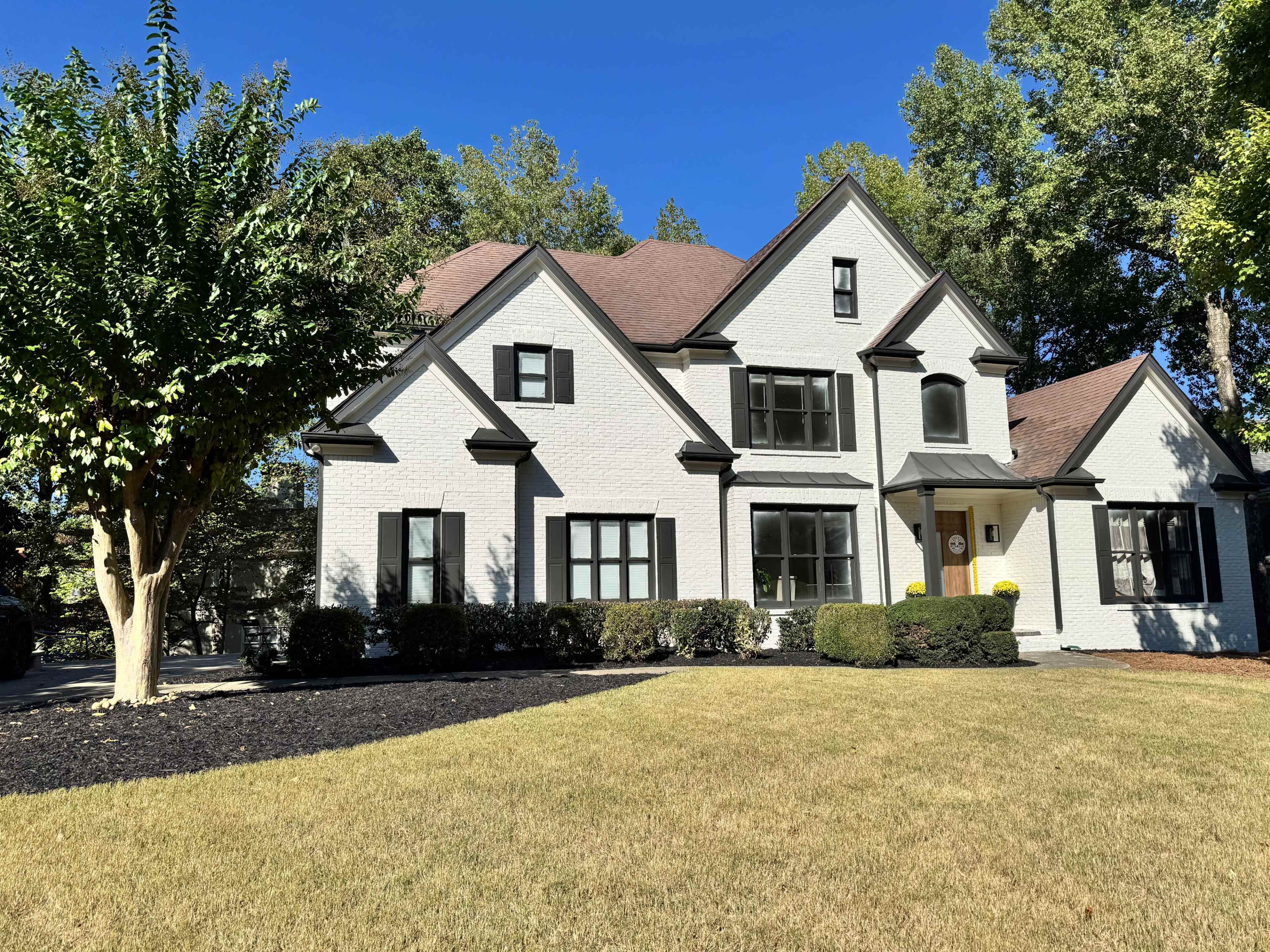 A two-story, white brick house with black window frames sits in a landscaped yard featuring grass and shrubs.