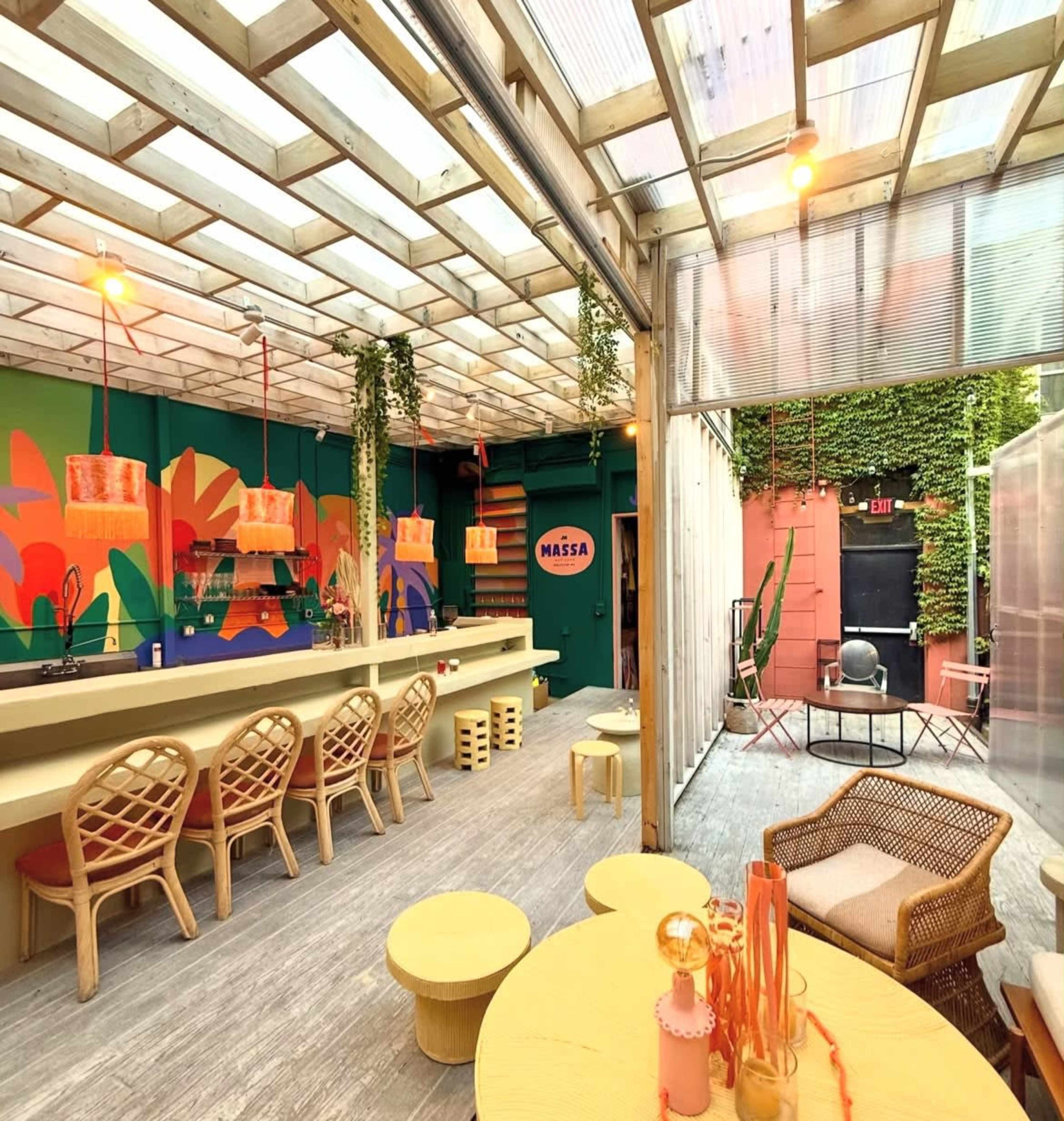 The image shows a modern indoor space with a bar, colorful wall art, and seating arrangements that include woven chairs and round tables.