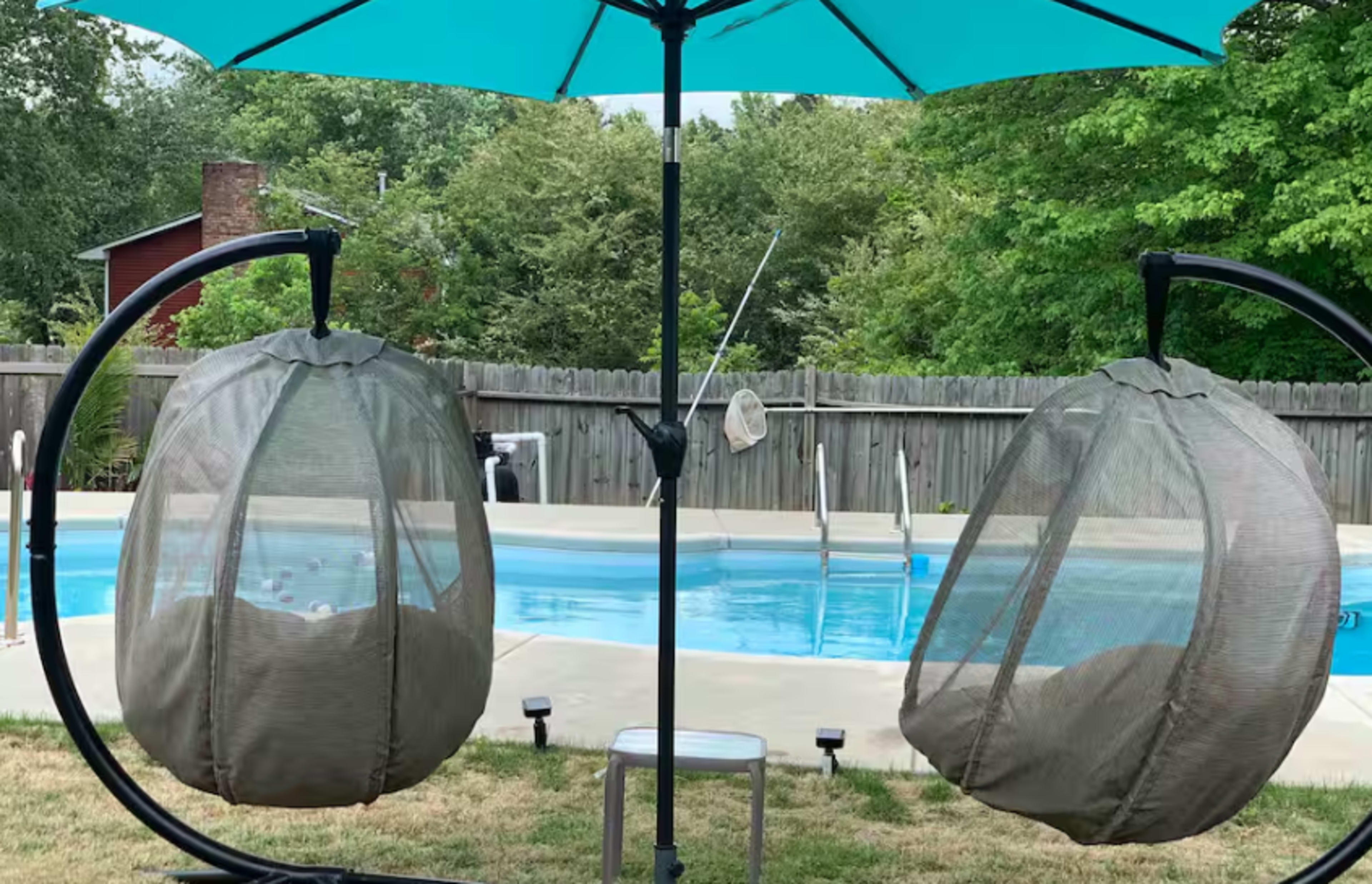 The image shows two hanging chairs covered with mesh fabric, suspended under a large turquoise umbrella, positioned next to a swimming pool surrounded by grass and trees.