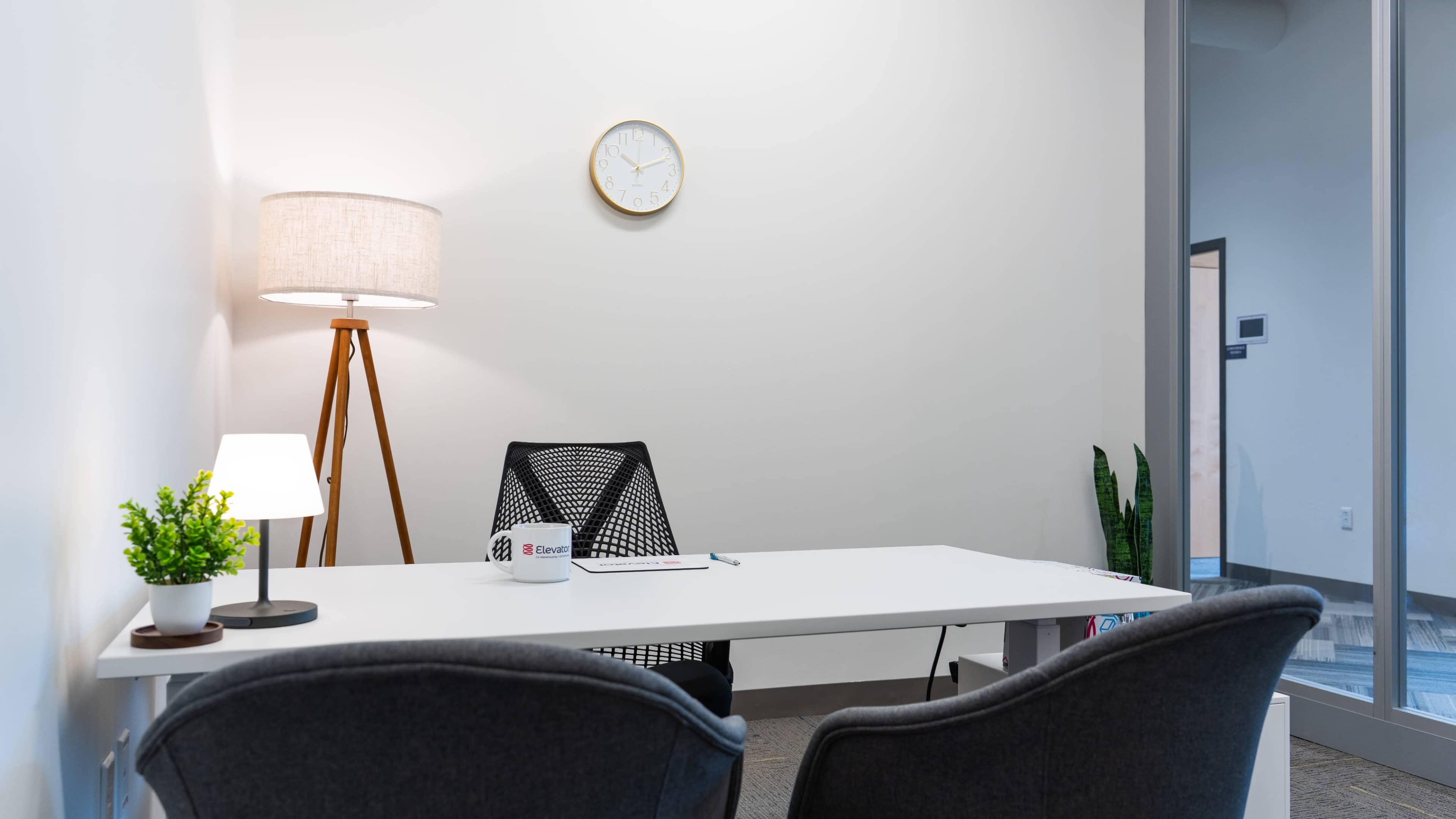 A modern office space features a white desk, two chairs, a lamp, and a clock on the wall, with a plant in the corner.
