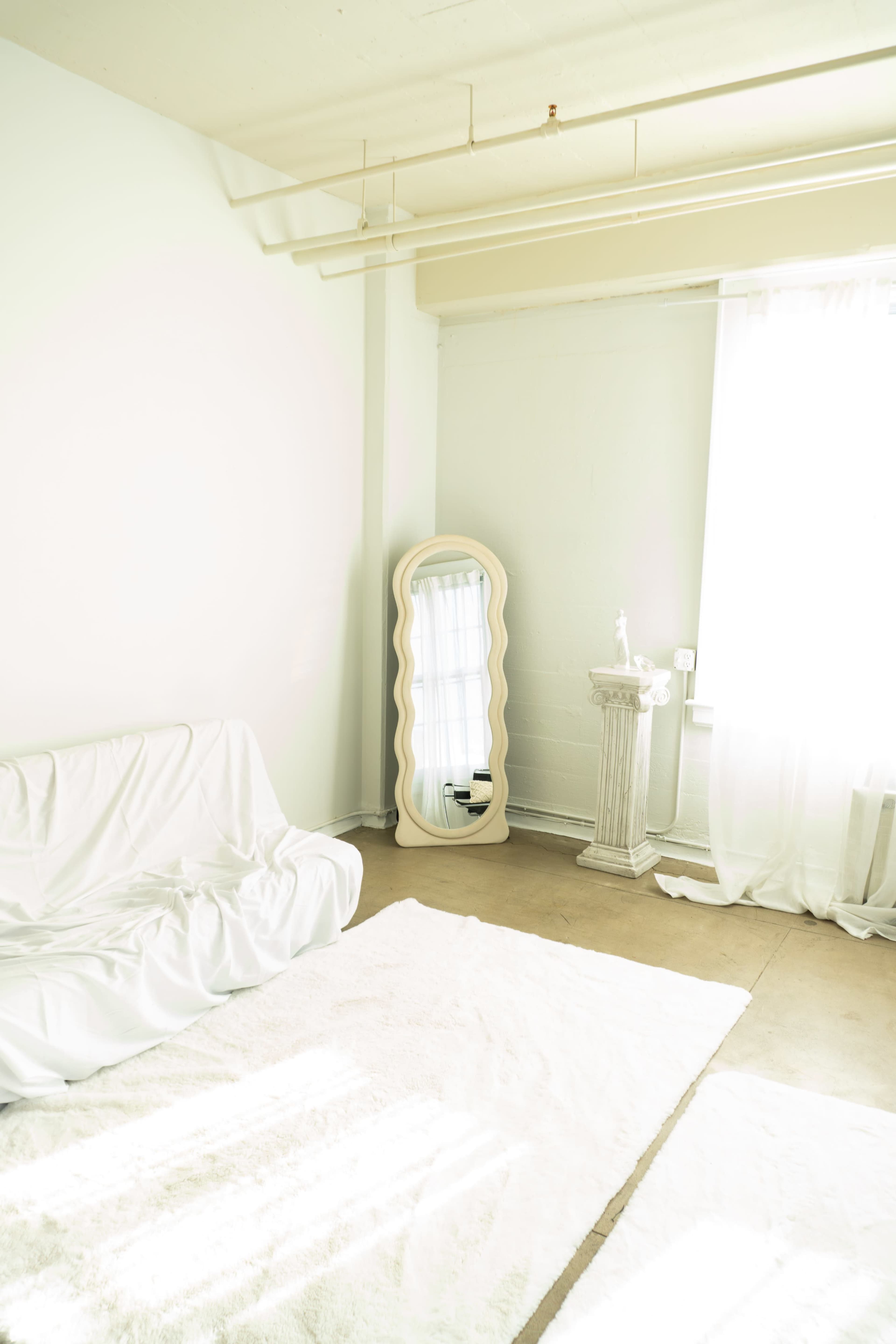 The room features a white couch, a large wavy mirror, and a decorative column beside a window covered with sheer curtains.