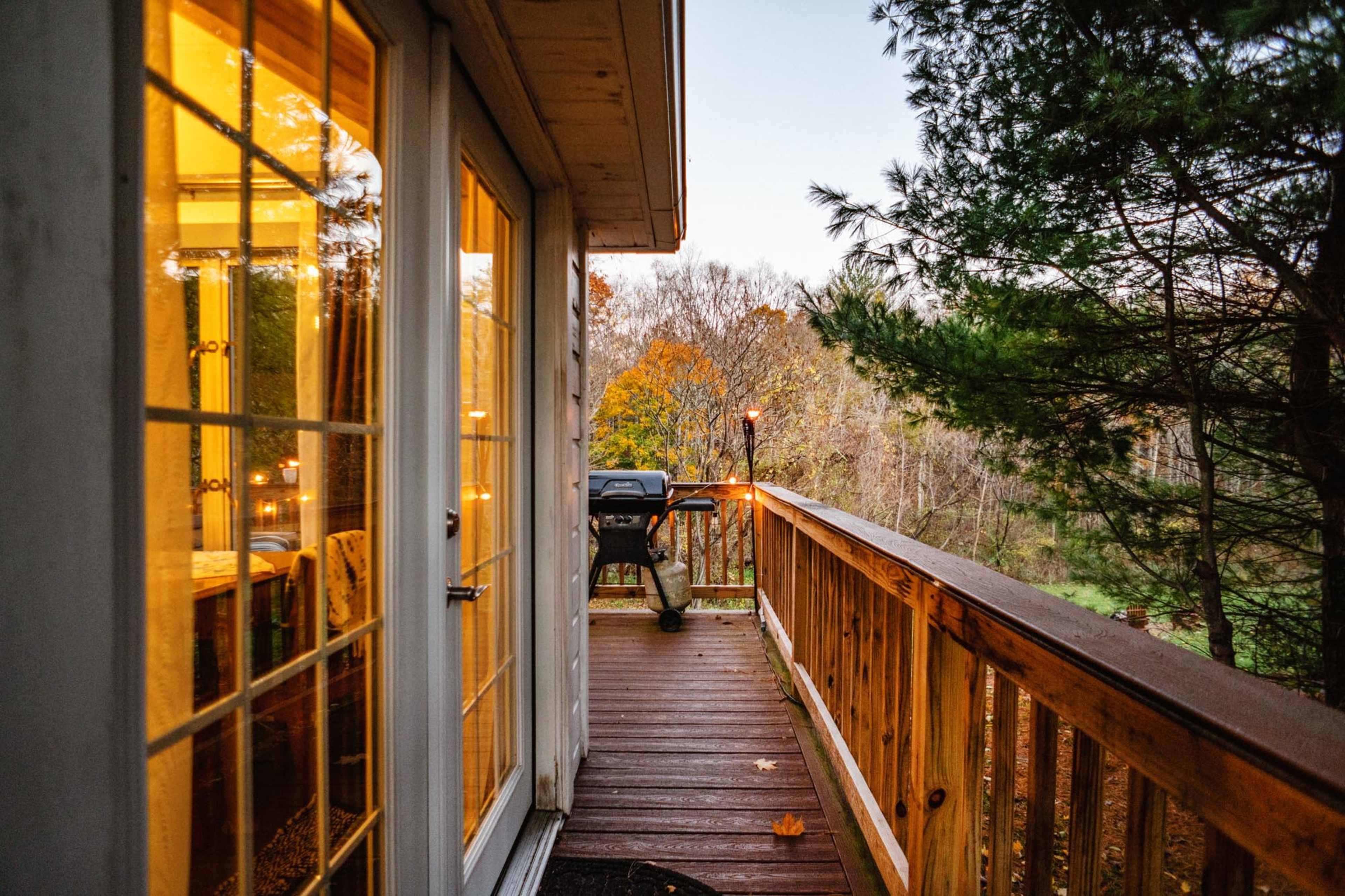 A wooden deck extends from a house, featuring a barbecue grill and surrounded by trees with fall foliage.