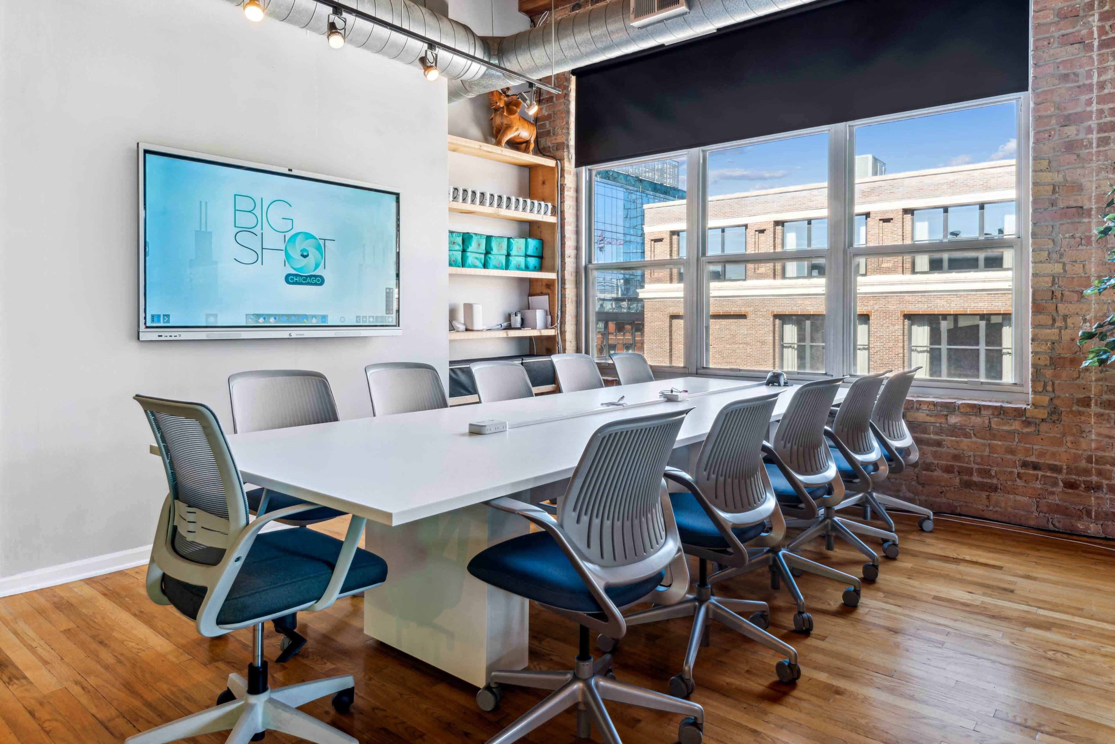 A modern conference room features a long white table surrounded by ergonomic chairs, with large windows providing natural light and a mounted screen displaying a company logo.