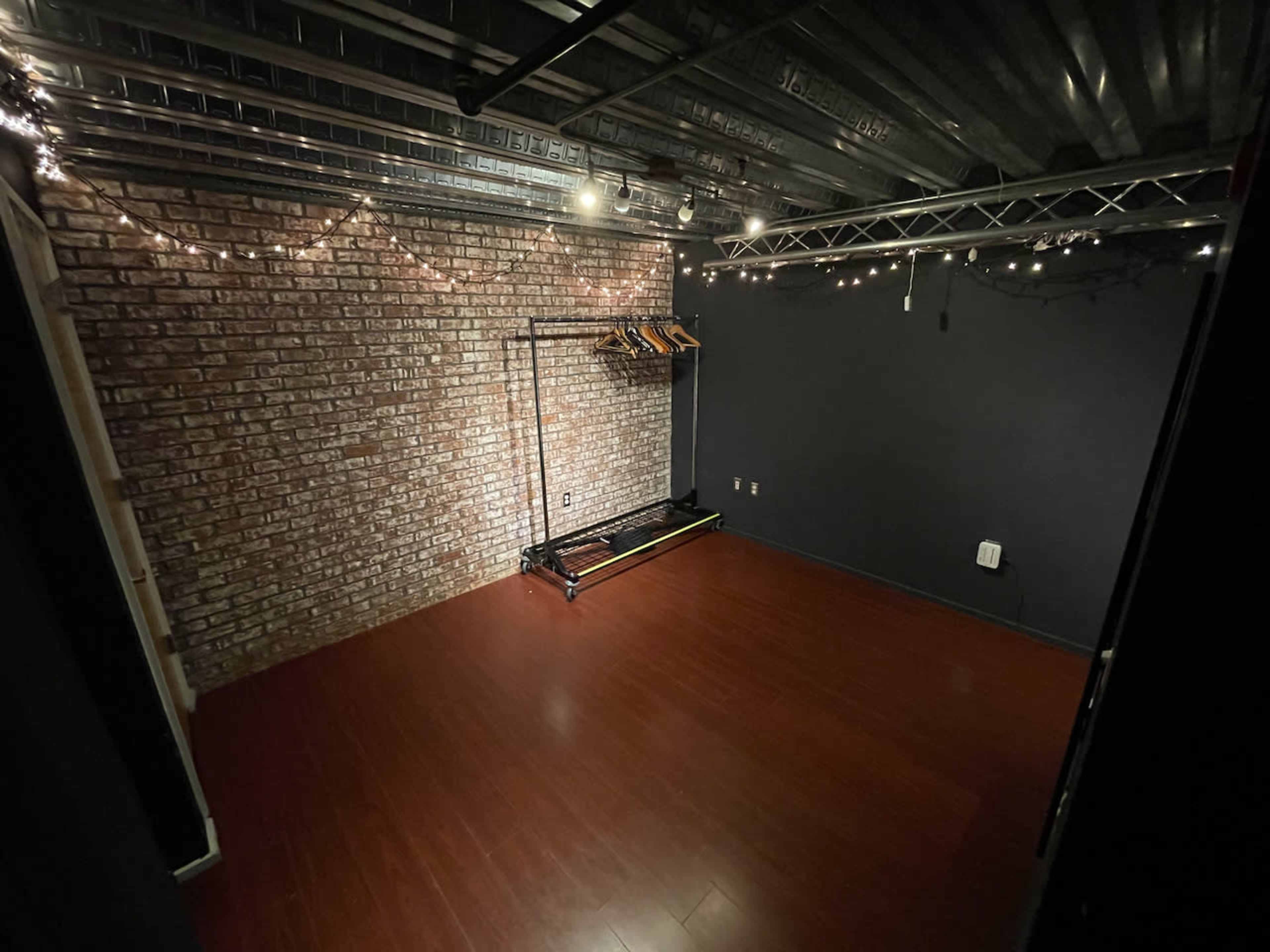 The image shows an empty room with a brick wall, dark-painted walls, a wooden floor, and a clothes rack against one wall, illuminated by string lights.