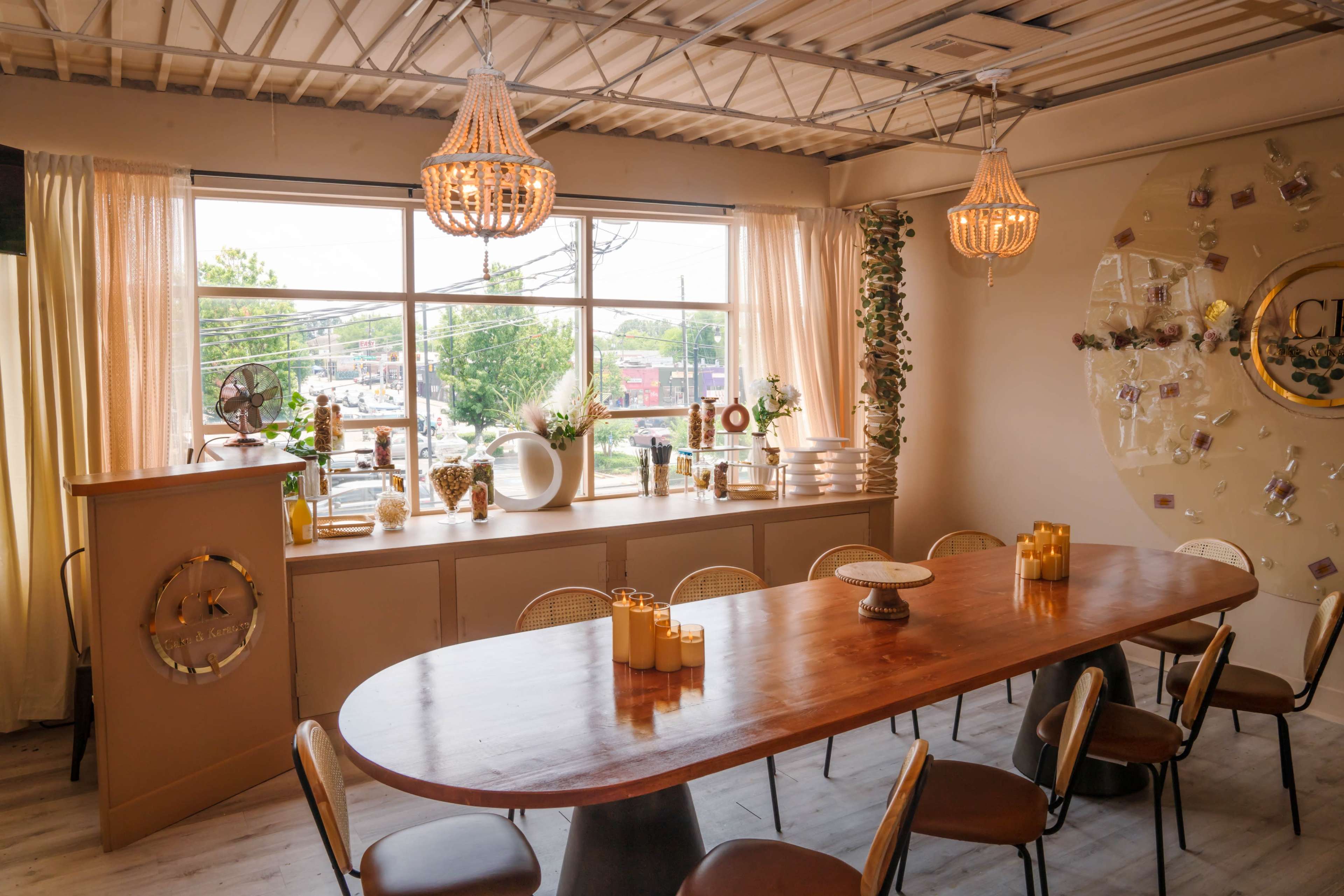 A spacious dining area features a long wooden table surrounded by chairs, with large windows allowing natural light to illuminate the decor and plants along the walls.