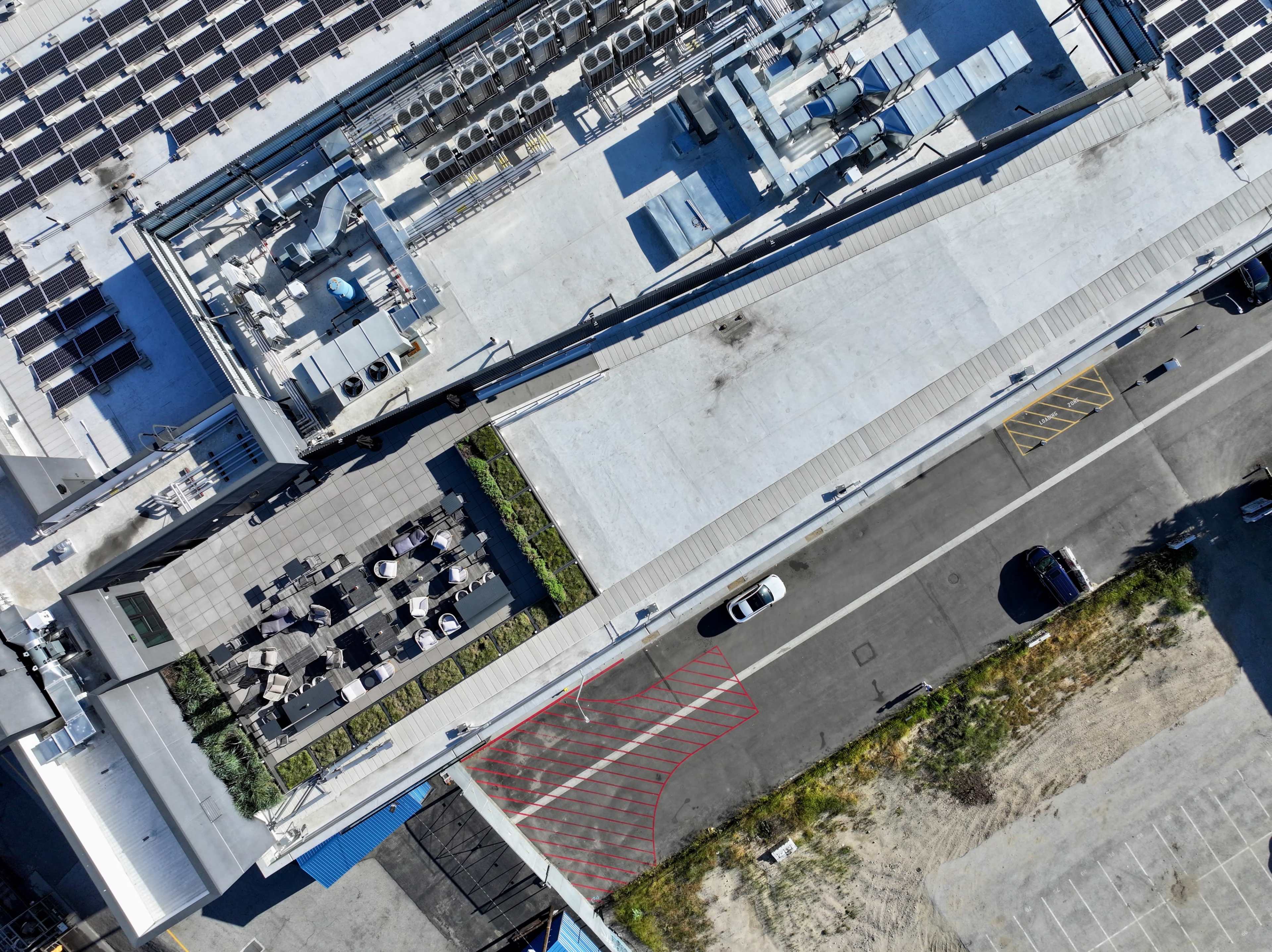 An overhead view of a commercial building featuring a rooftop area with outdoor seating and solar panels, surrounded by various mechanical units and an adjacent parking lot.