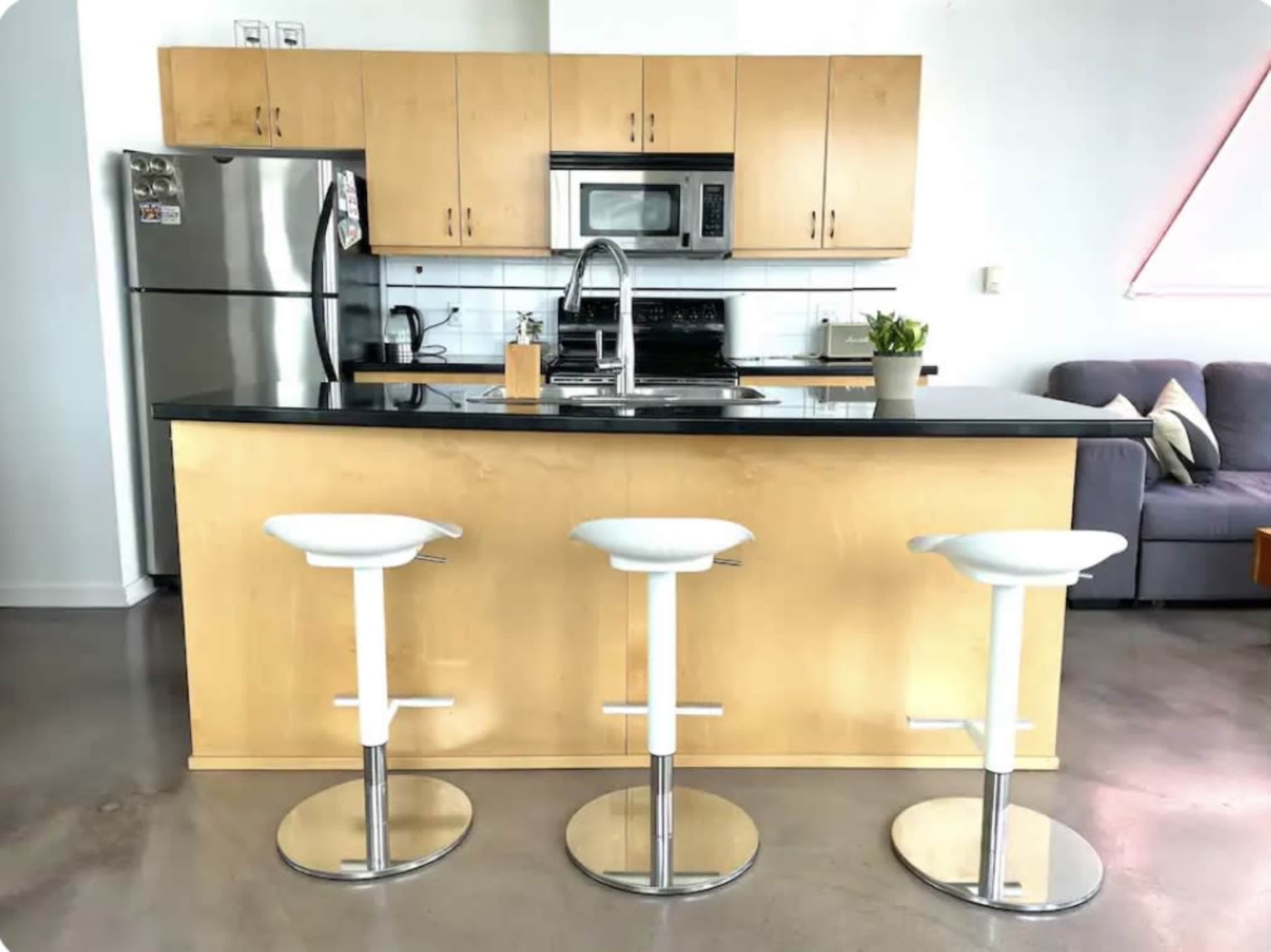 A modern kitchen featuring a central island with three white bar stools, stainless steel appliances, and wooden cabinetry.