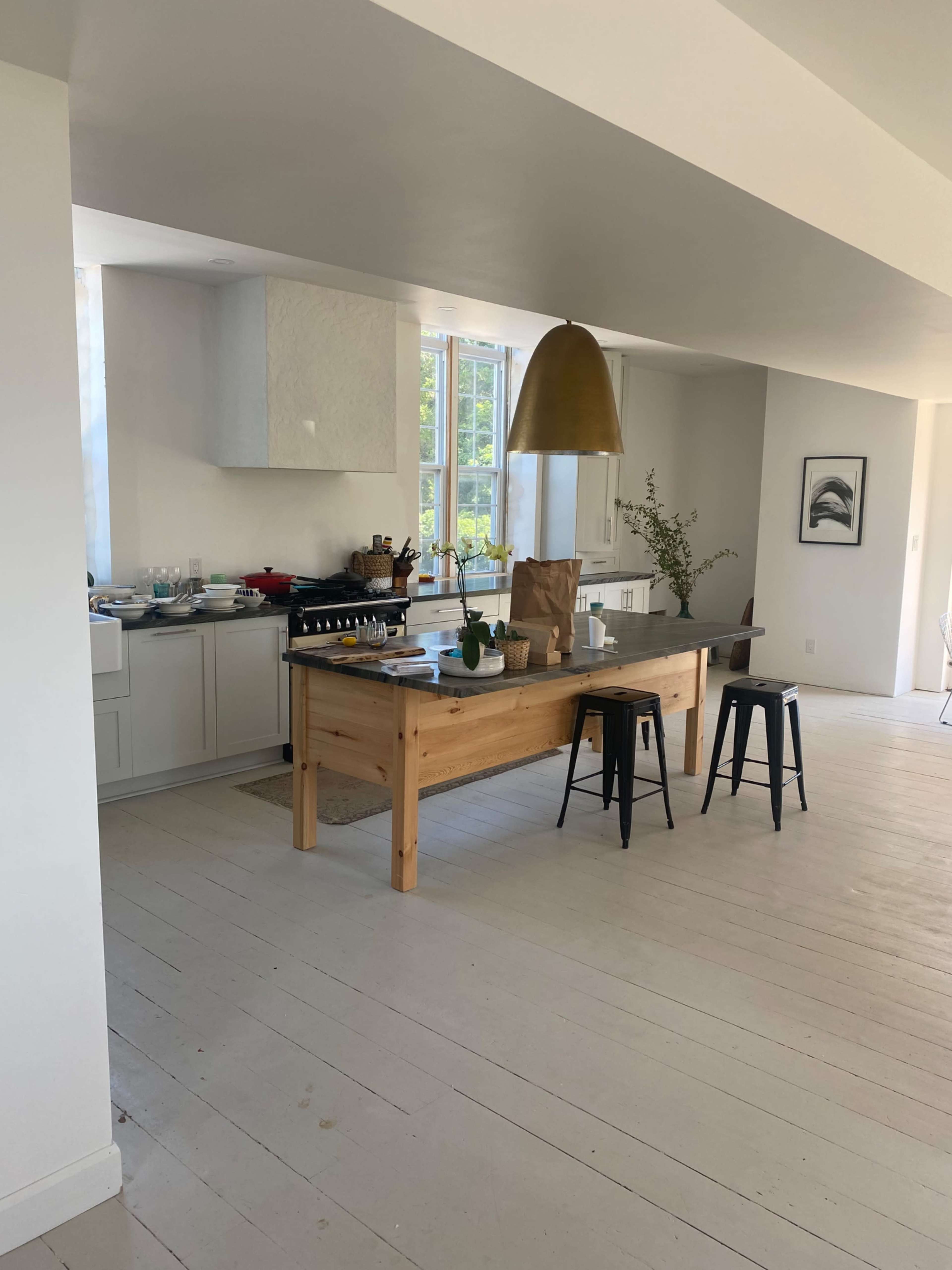 A modern kitchen features a wooden table with black stools, a gold pendant light overhead, and a clean, open design with white walls.