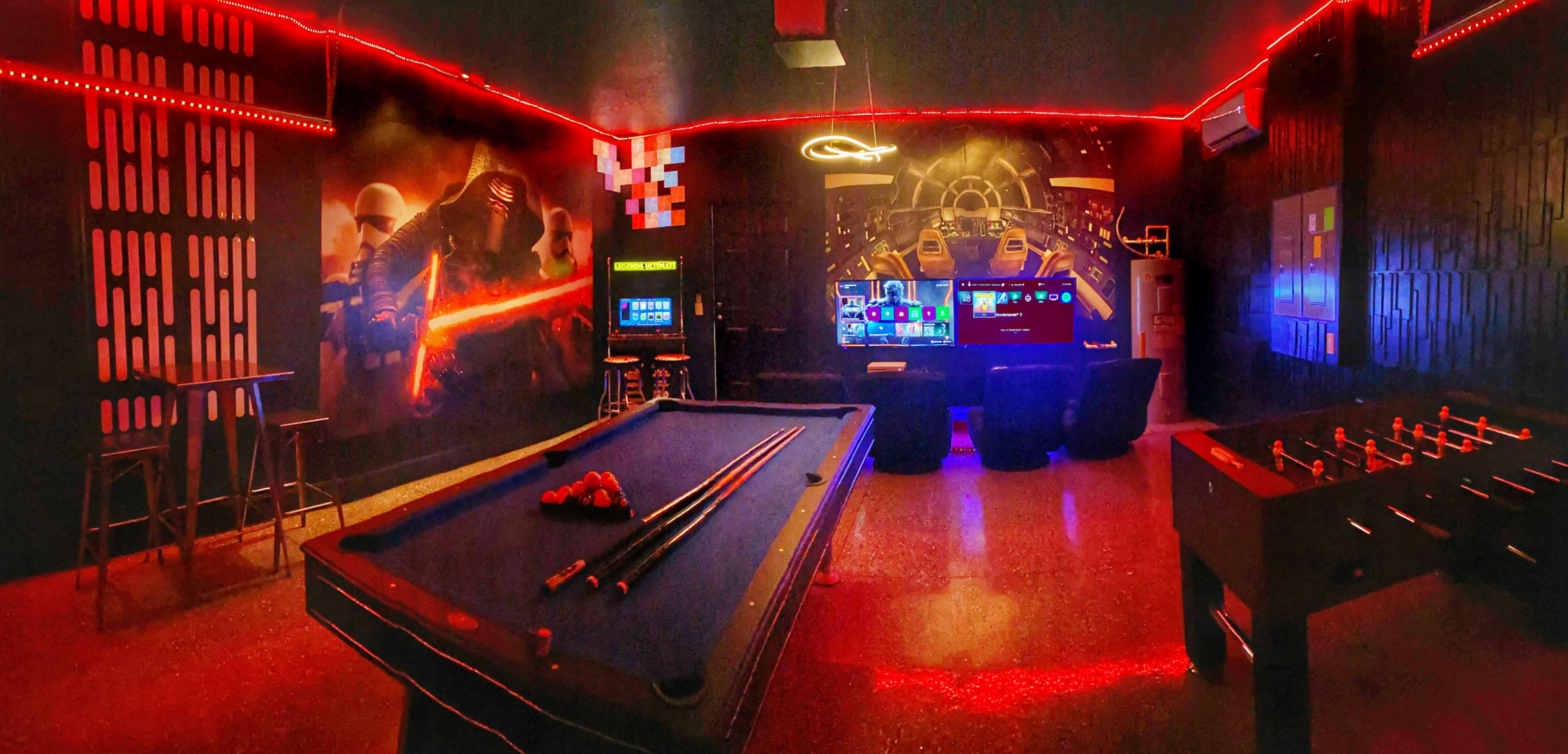 A dimly lit game room featuring a pool table, foosball table, arcade machines, and vibrant red neon lighting.