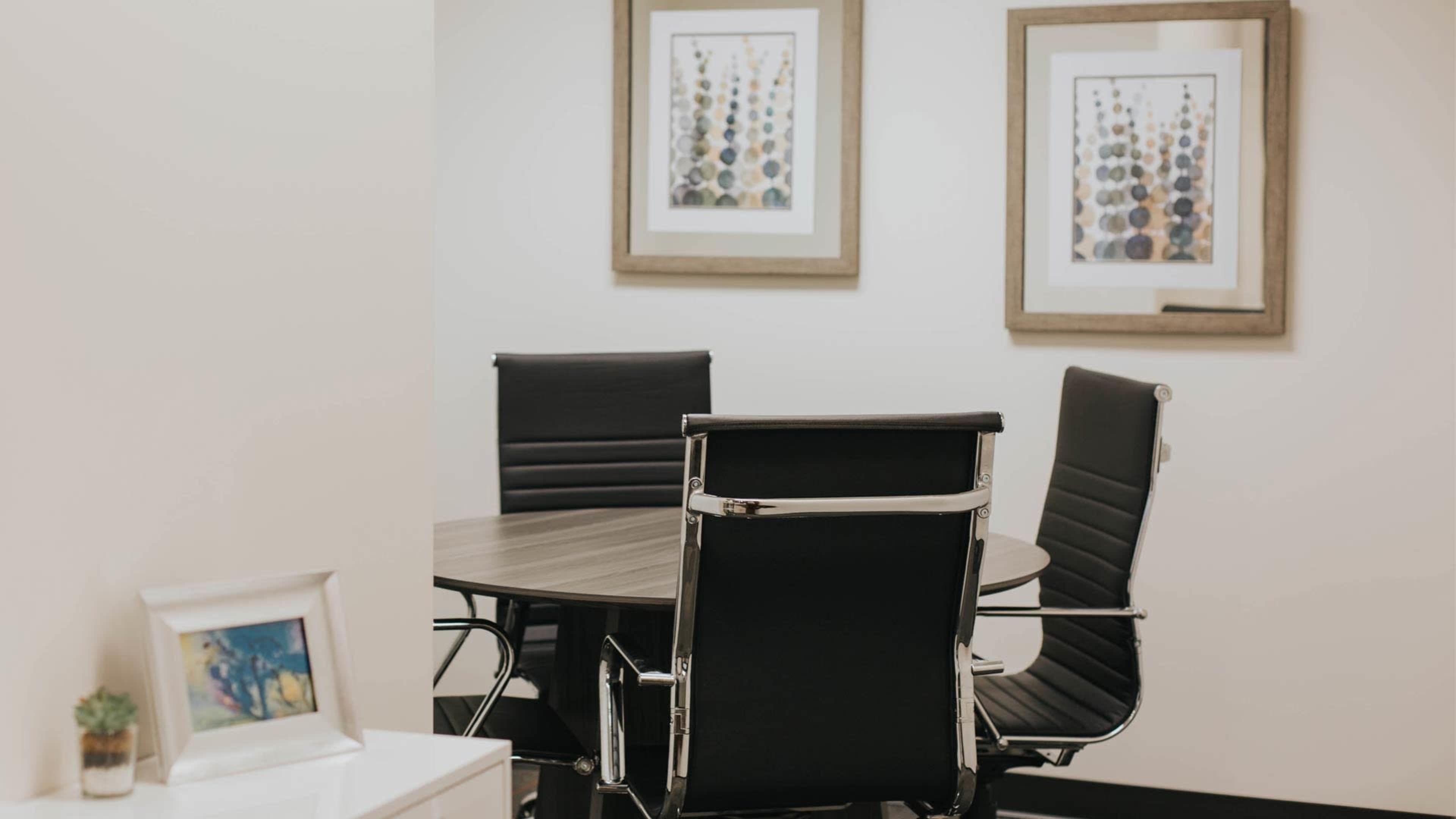 A small meeting room features a round table surrounded by four black chairs, with framed artwork on the walls.