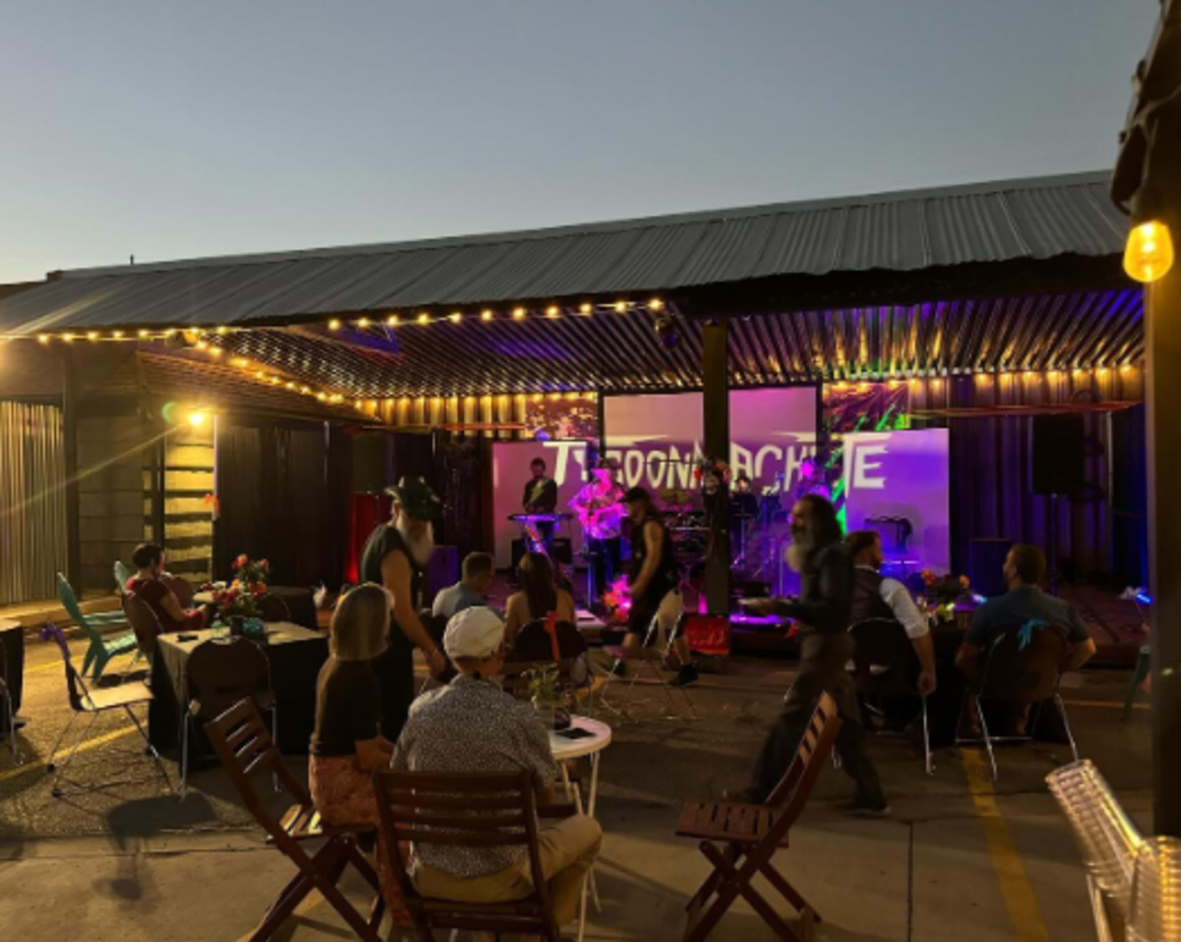 A live band performs on a lit outdoor stage while attendees enjoy food and drinks at tables nearby.