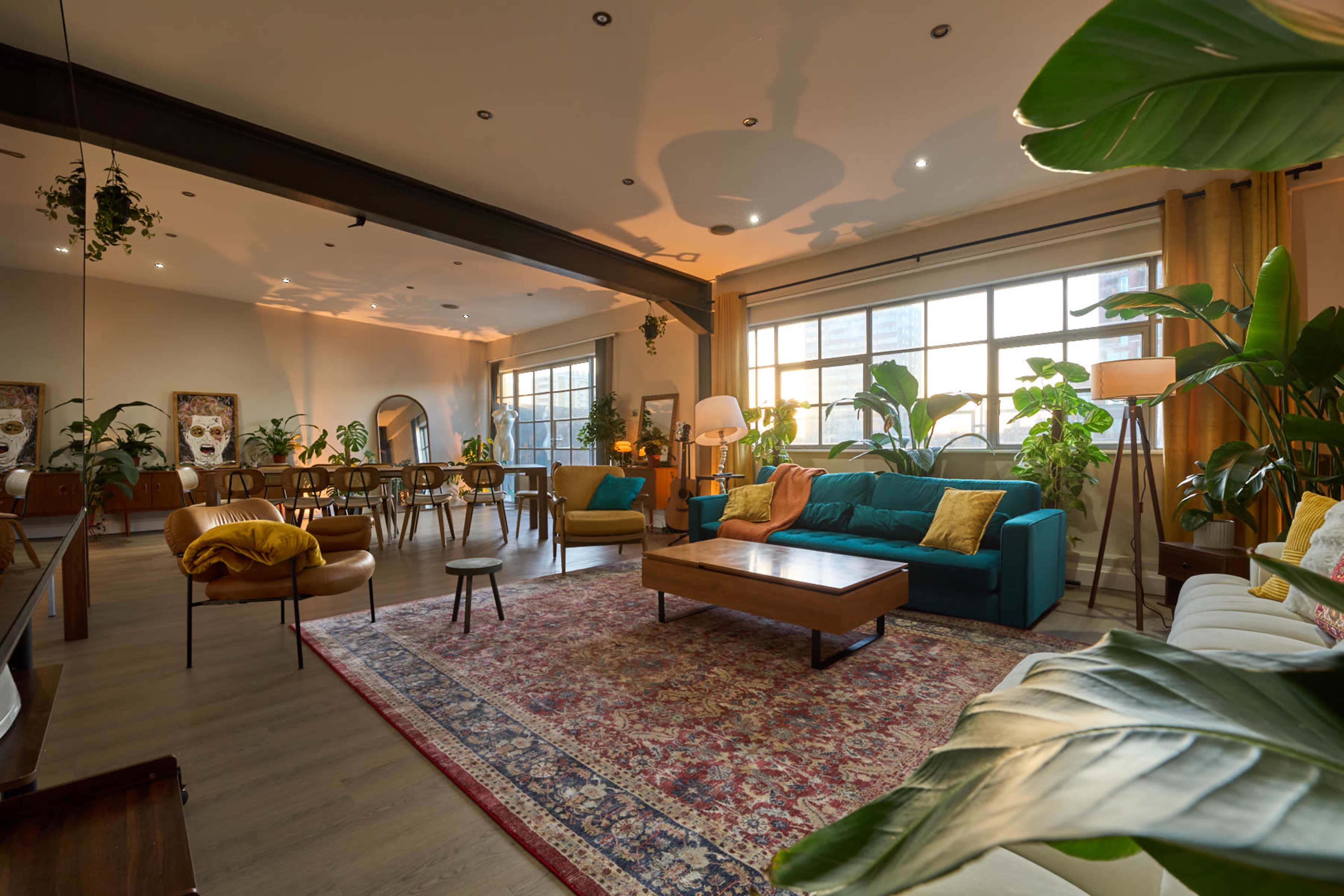 The image shows a spacious living area featuring teal and white sofas, a large wooden coffee table, potted plants, and a dining table in the background, all illuminated by natural light coming through large windows.