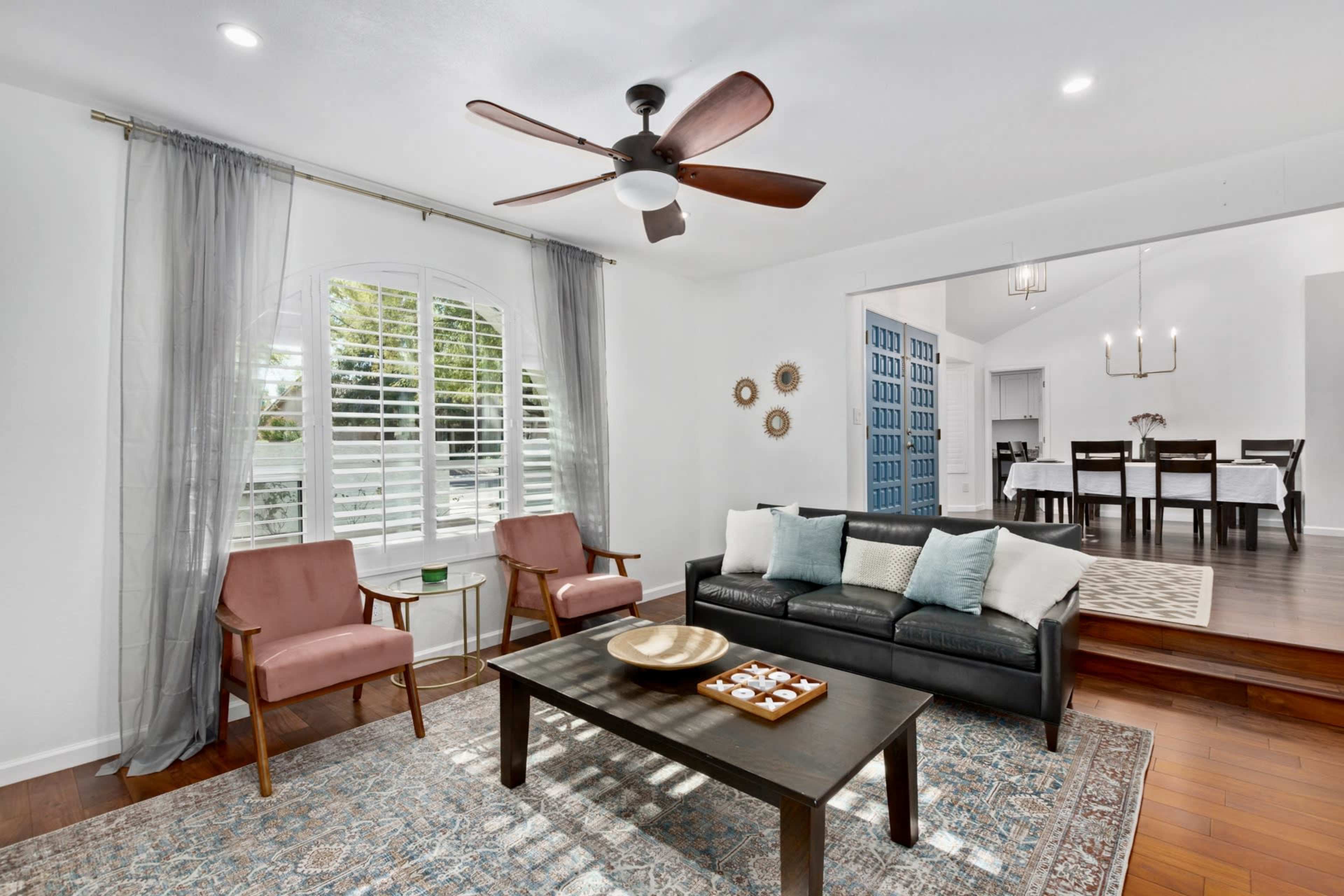 The image shows a bright living room with two armchairs, a black sofa, a coffee table, and a view of a dining area in the background.