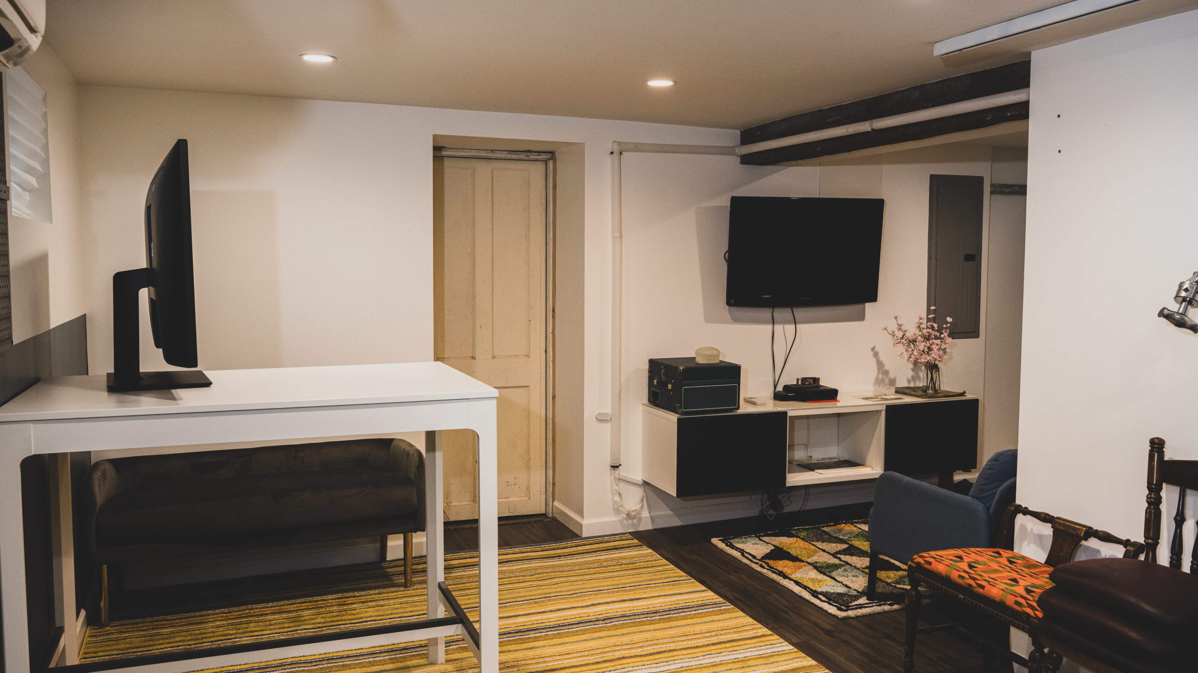 A basement room features a television mounted on the wall, a small sofa, a table, and a colorful rug.