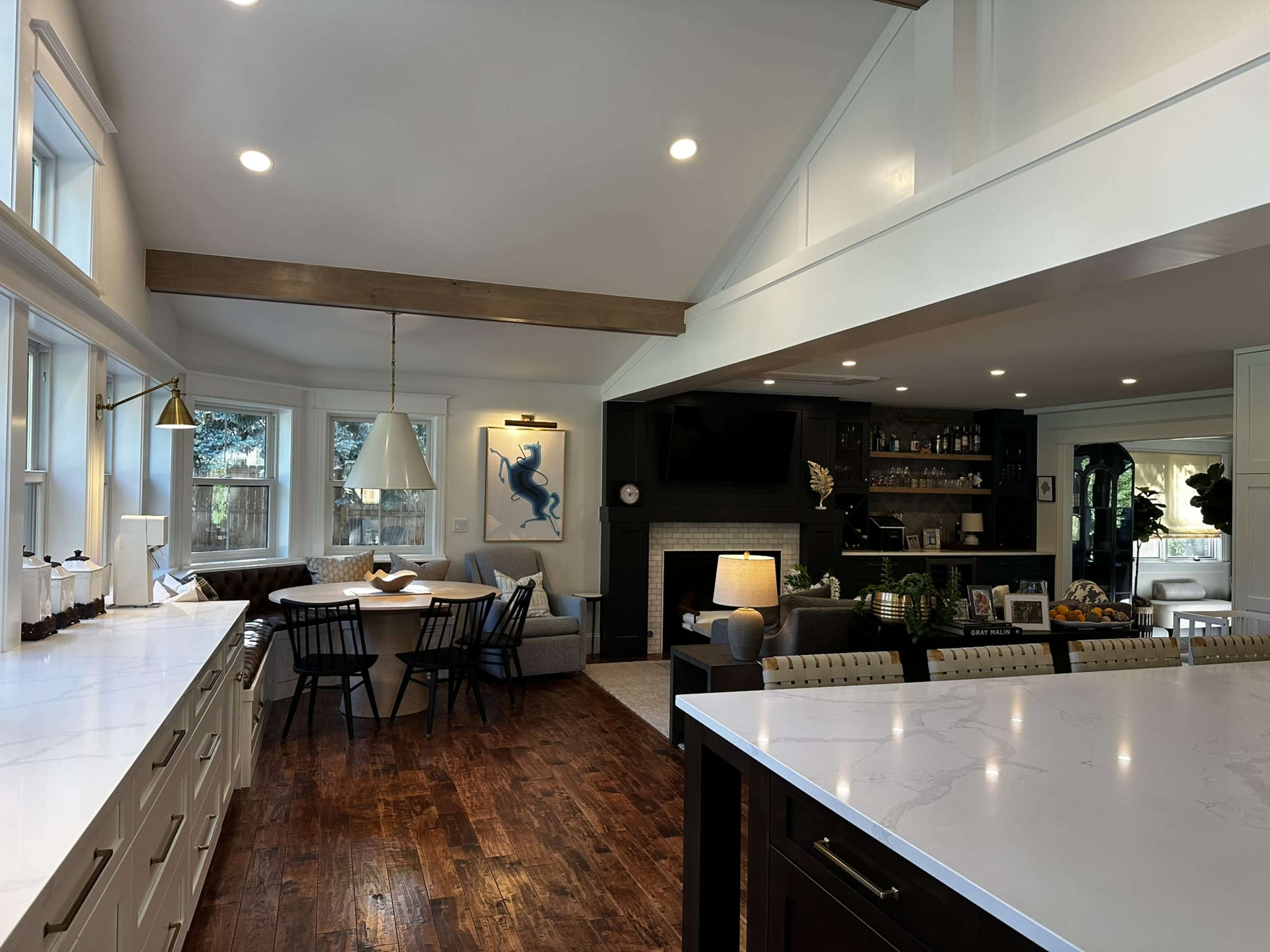 The image shows a modern kitchen with an open layout, featuring white cabinetry, a large island, and a dining area with a round table adjacent to a sitting area with a fireplace.