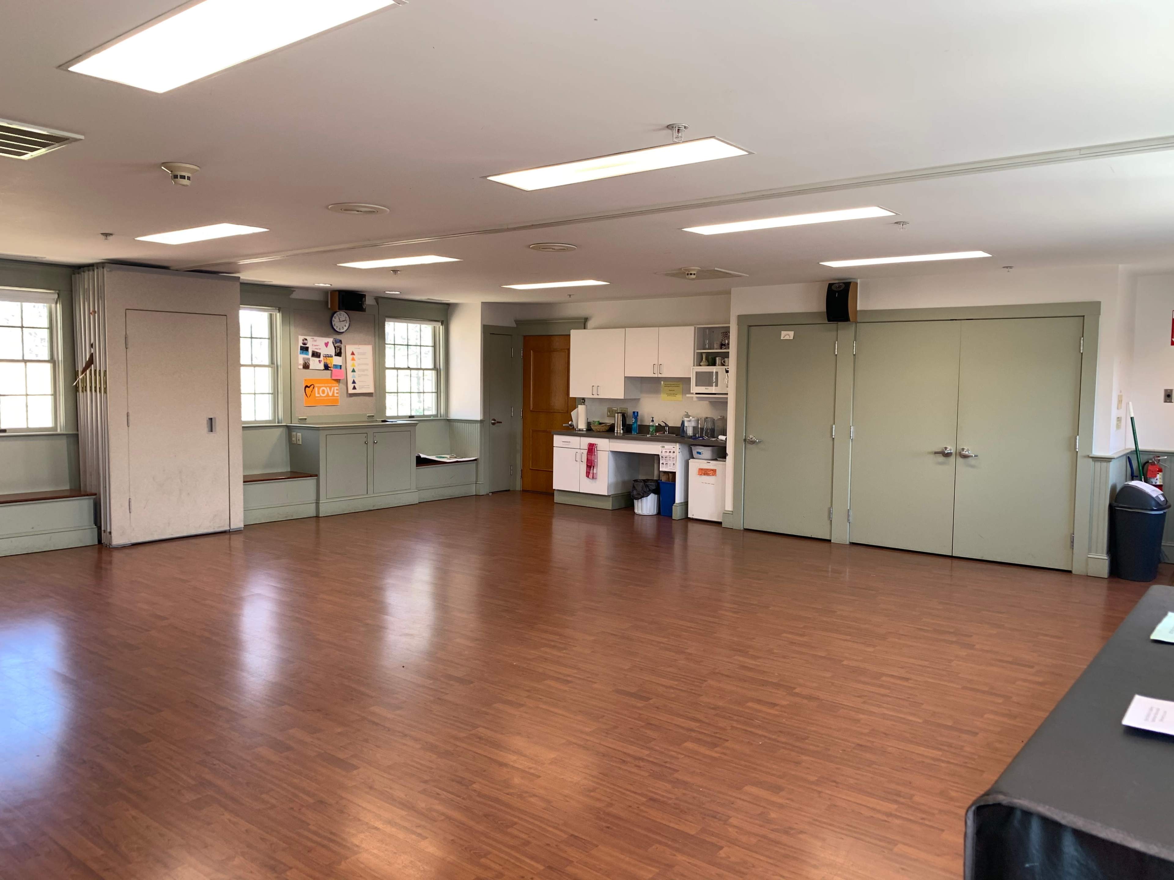 An empty room with wooden floors, bright overhead lights, a kitchenette area, and multiple doors along the walls.