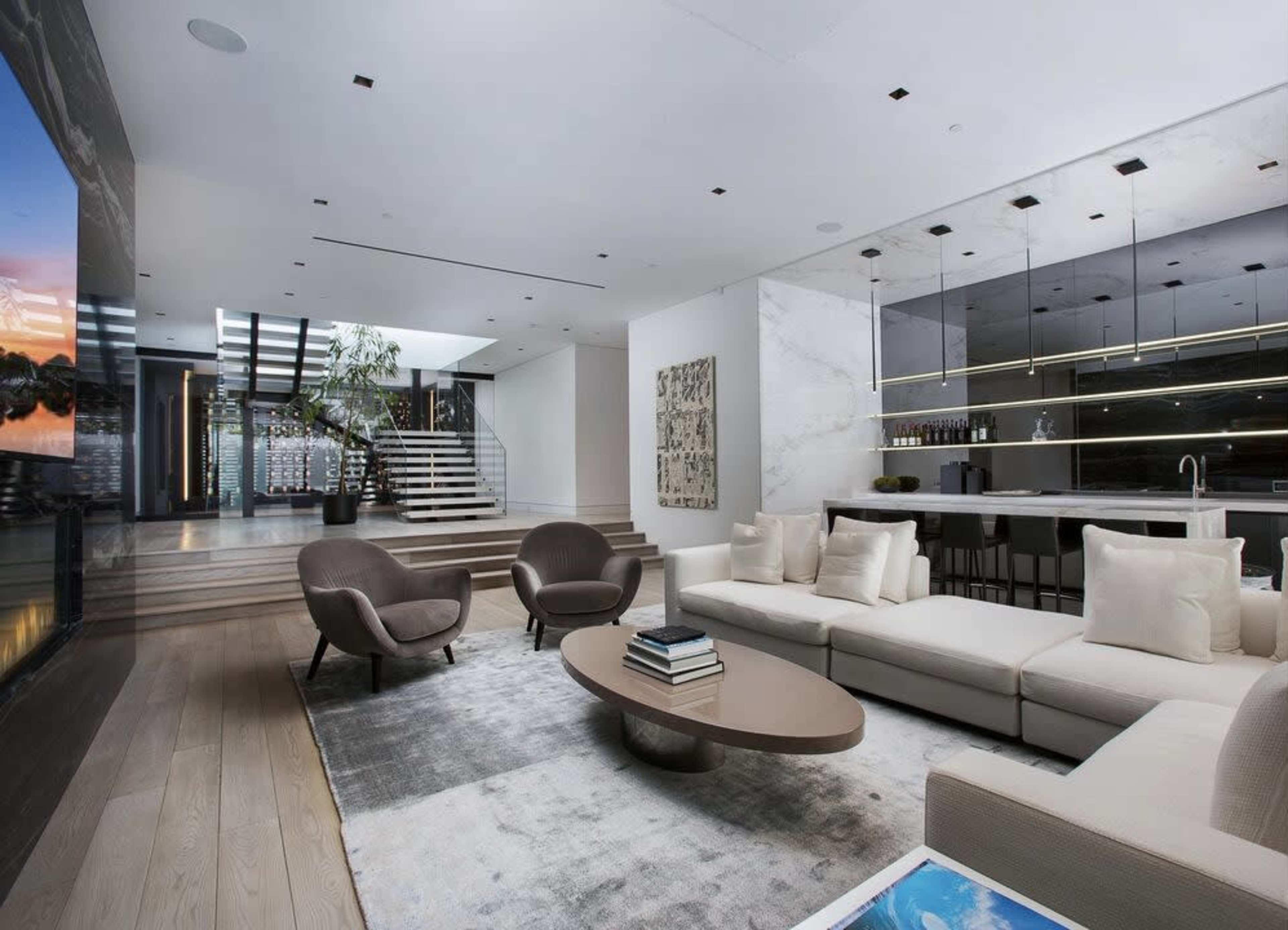 The image shows a modern living room with a sectional sofa, two armchairs, a coffee table, and a large window overlooking a staircase and an open kitchen area.