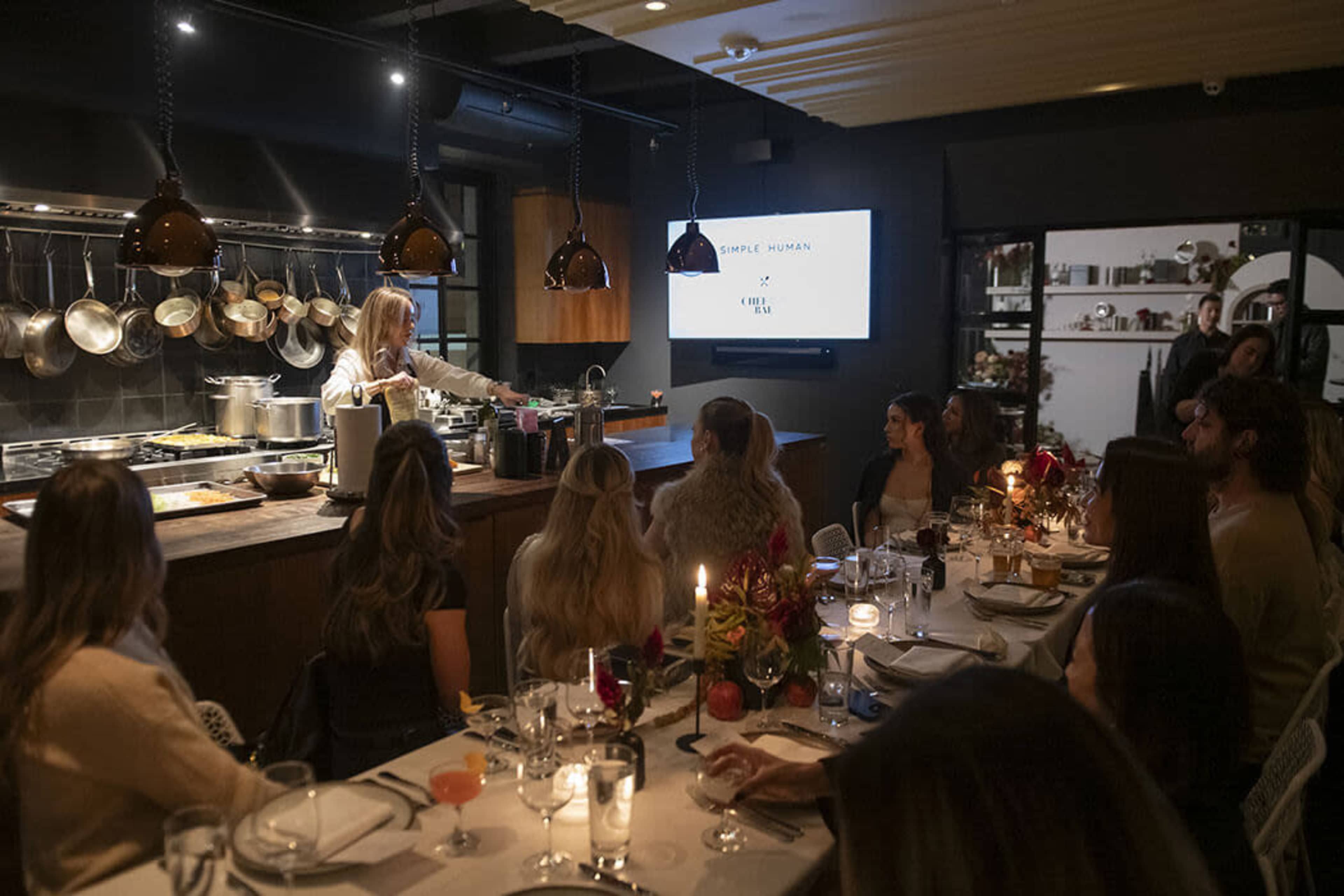 A chef presents a cooking demonstration to an audience seated at a long table in a cozy kitchen restaurant.