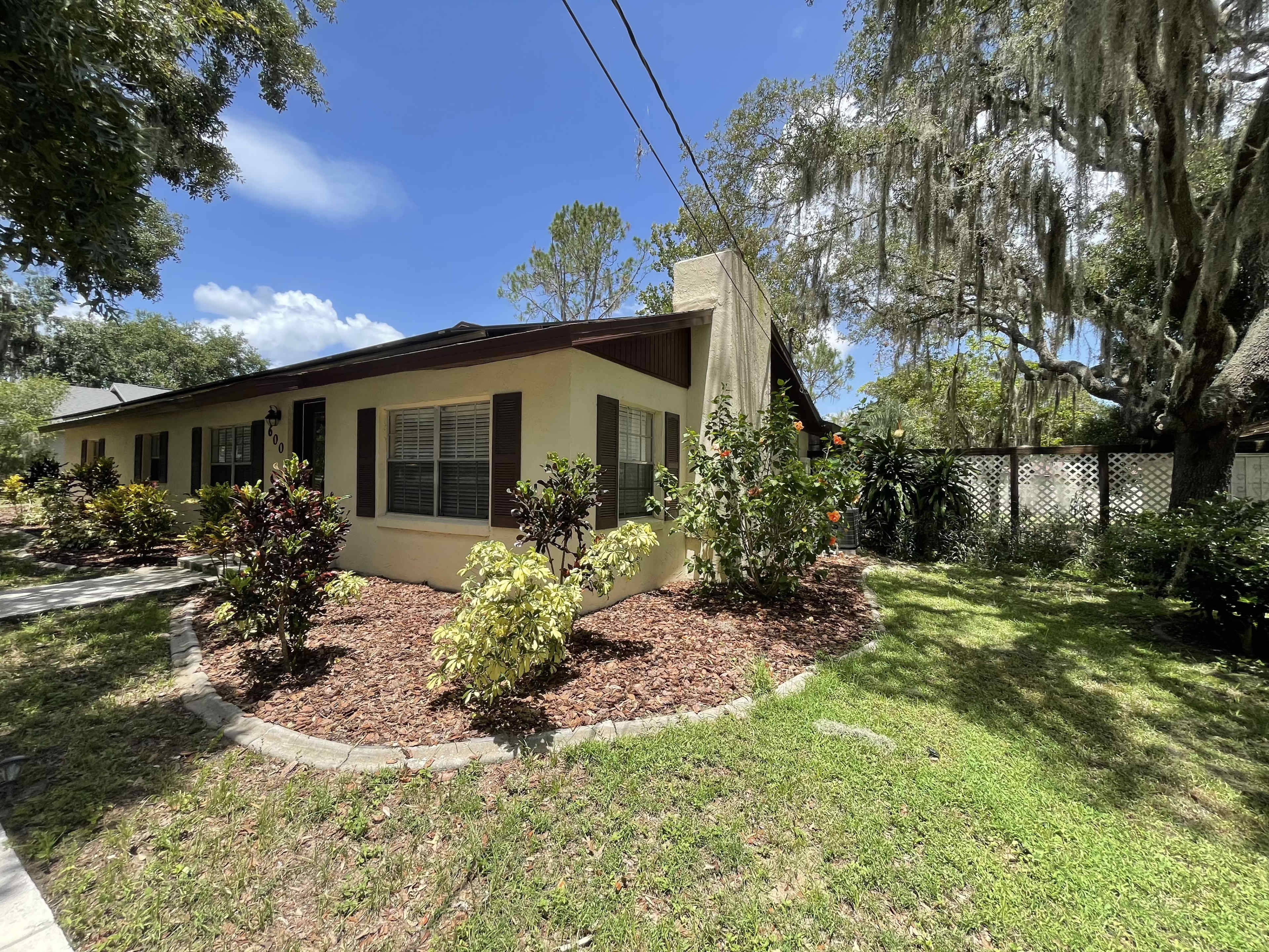 A single-story yellow house with brown shutters is surrounded by landscaped gardens and trees in a residential area.