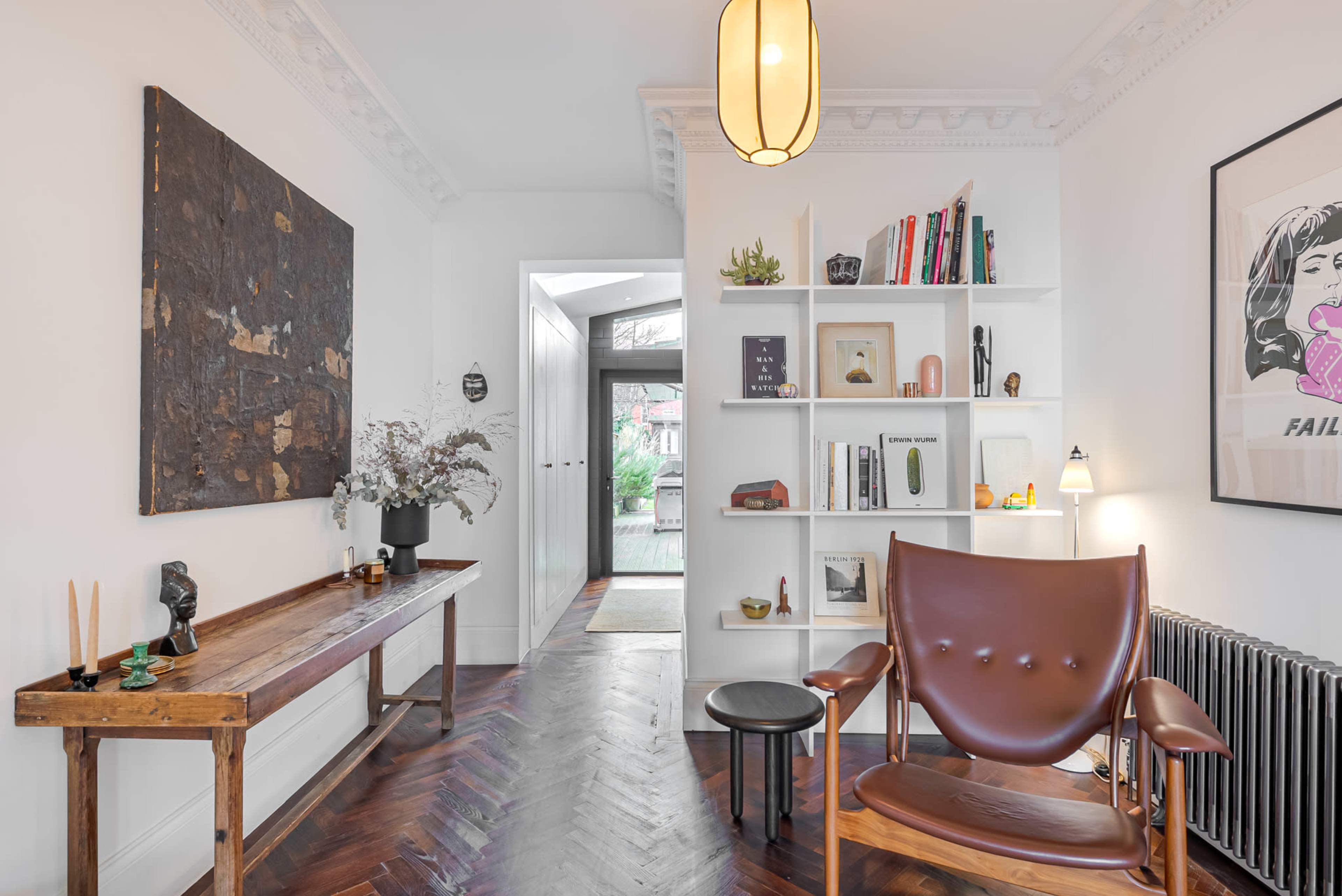 The image shows a well-lit hallway featuring a wooden bench, a bookshelf filled with various items, and a leather armchair next to a small side table.