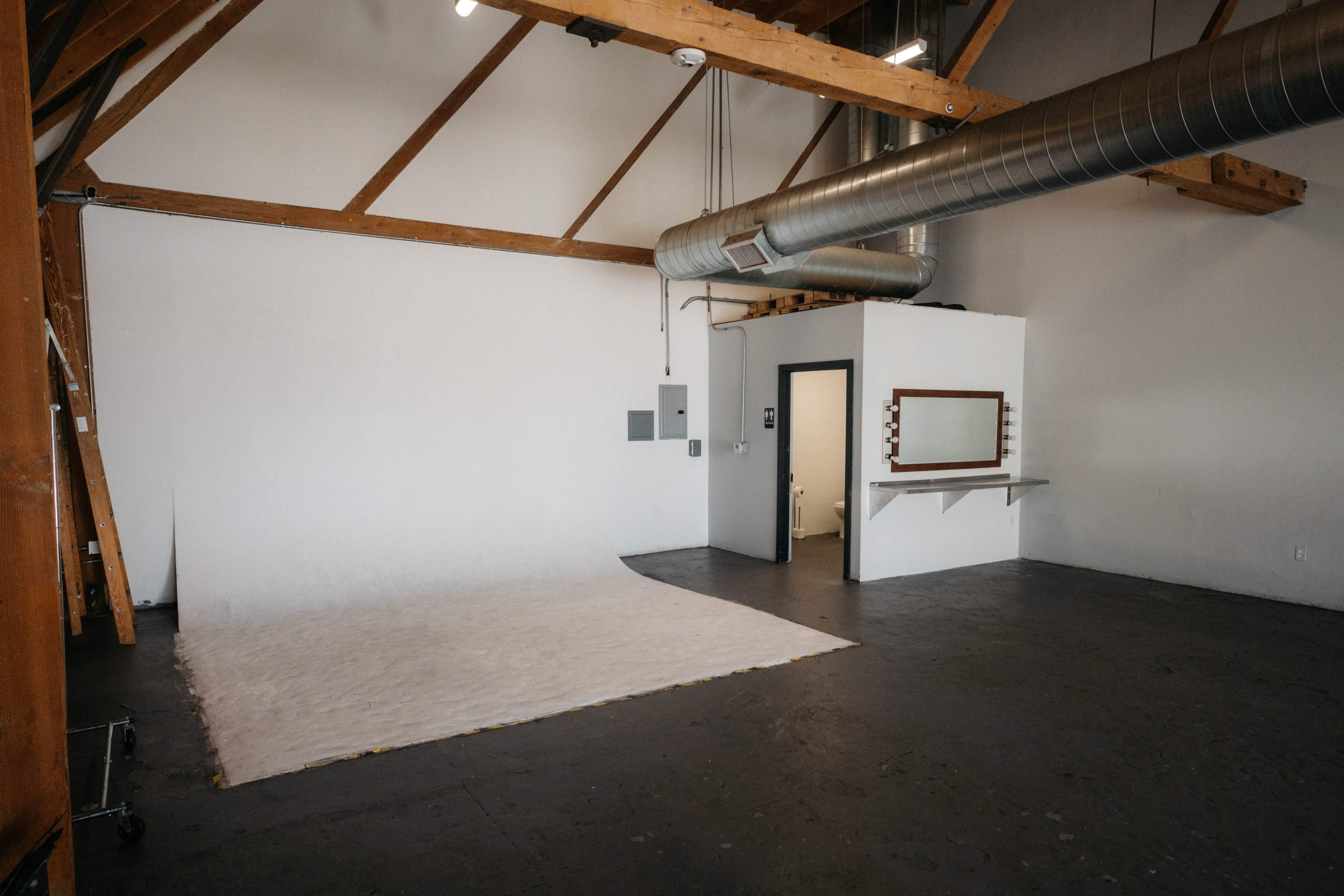The image shows a spacious, empty studio with wooden beams, a white backdrop, and a small mirror setup on a wall.