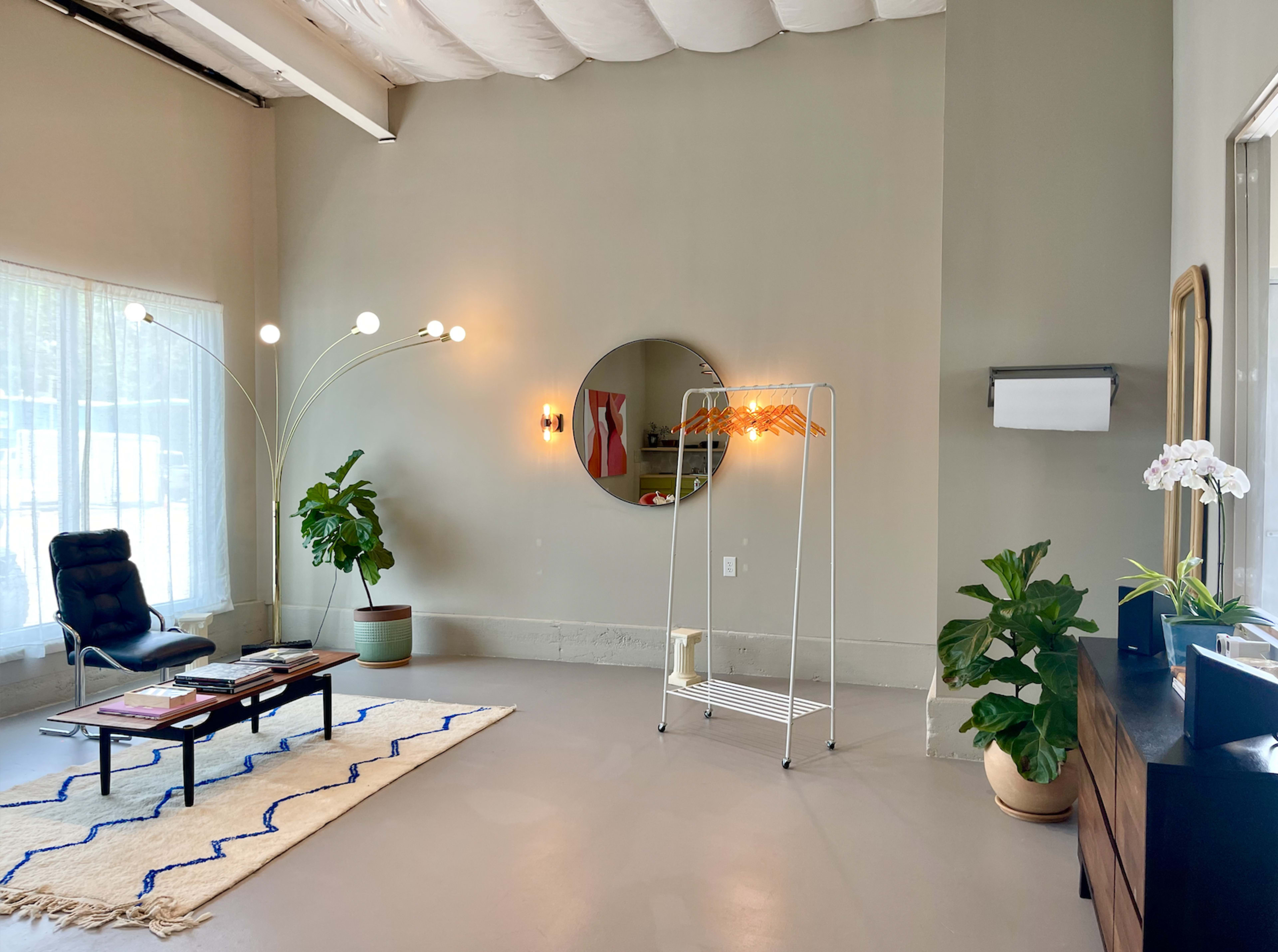 A modern interior living space with a black lounge chair, a round mirror, a clothing rack, a side table, and potted plants, all set against a neutral wall.