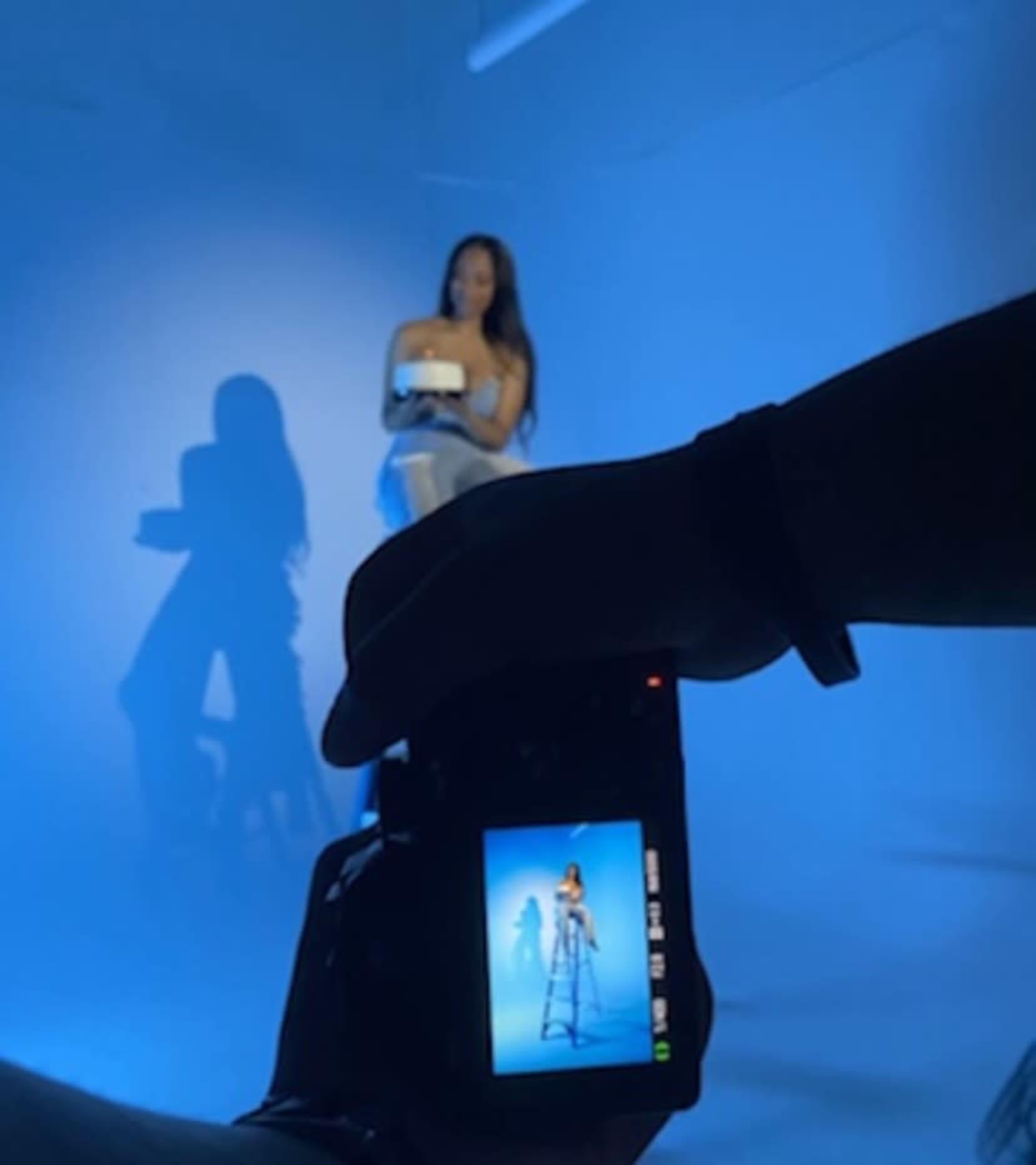 A person holds a camera, capturing a photo of a model seated on a stool in a blue-lit studio.
