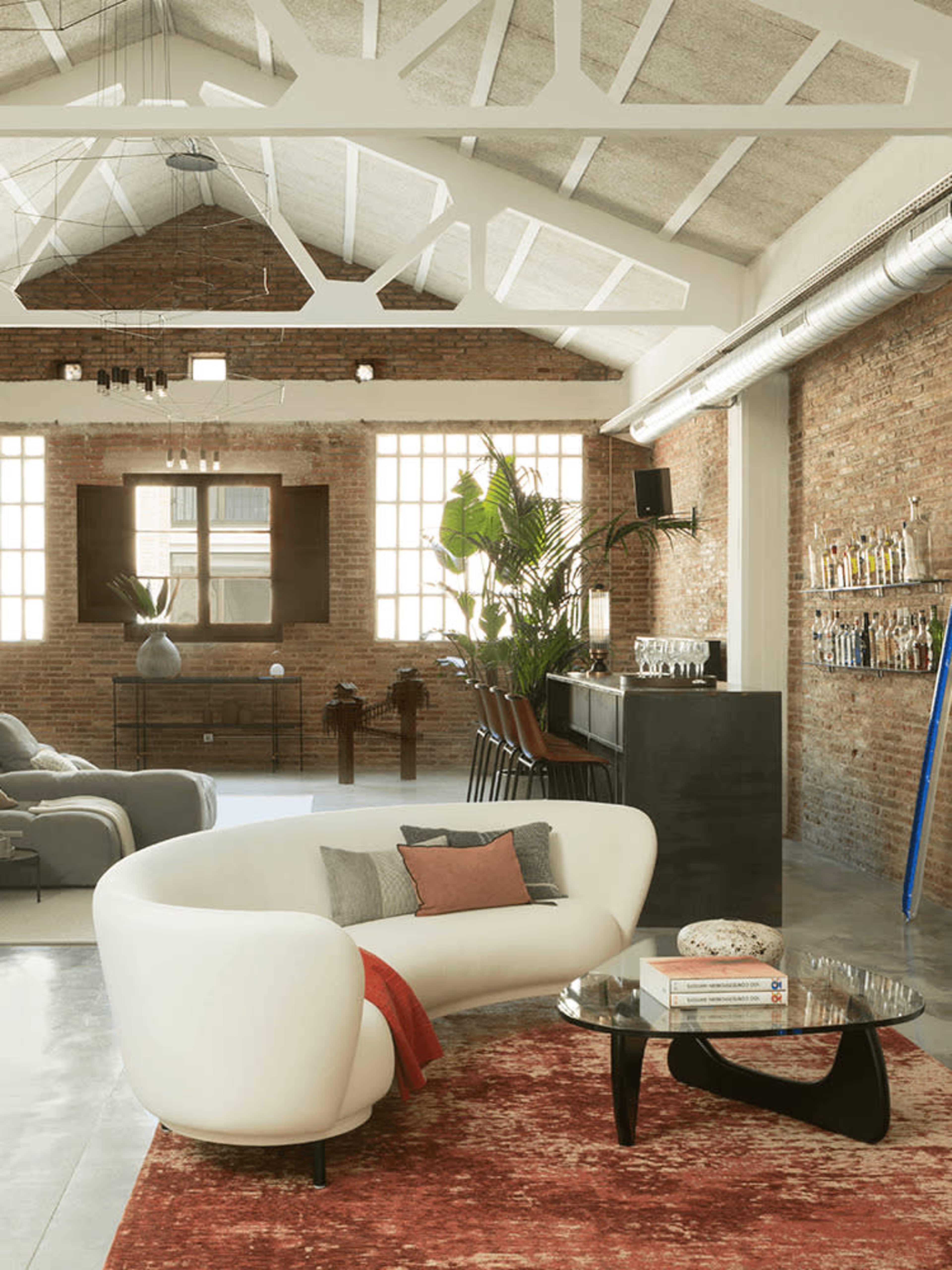 The image shows a modern living space with a curved white sofa, a glass coffee table, and exposed brick walls under a high ceiling.