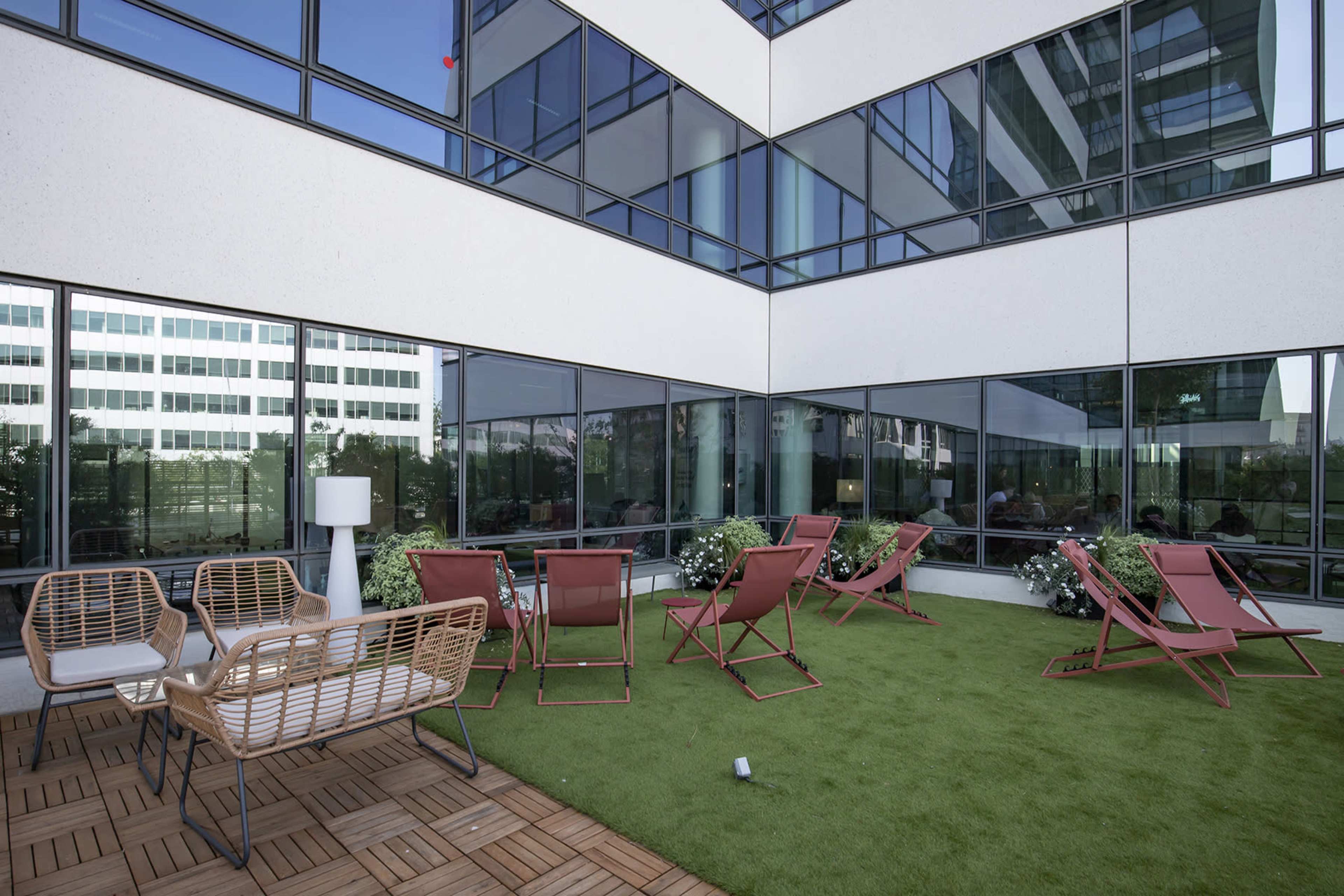 The image shows a rooftop terrace with green grass, several lounge chairs, a small table, and flowering plants surrounded by modern building windows.