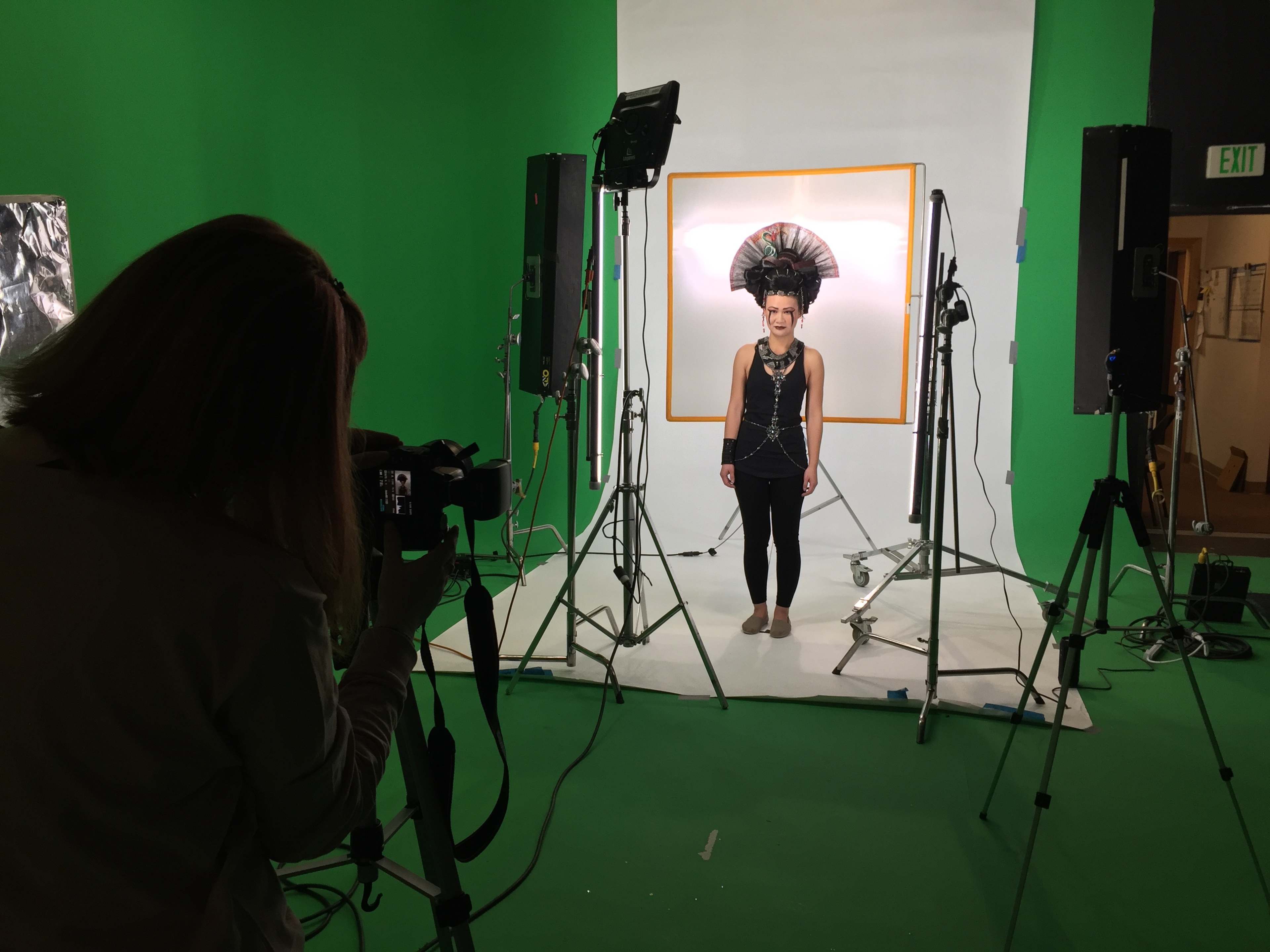 A person wearing a large feathered headdress poses for a photo in a studio with a green backdrop while another person takes the picture.