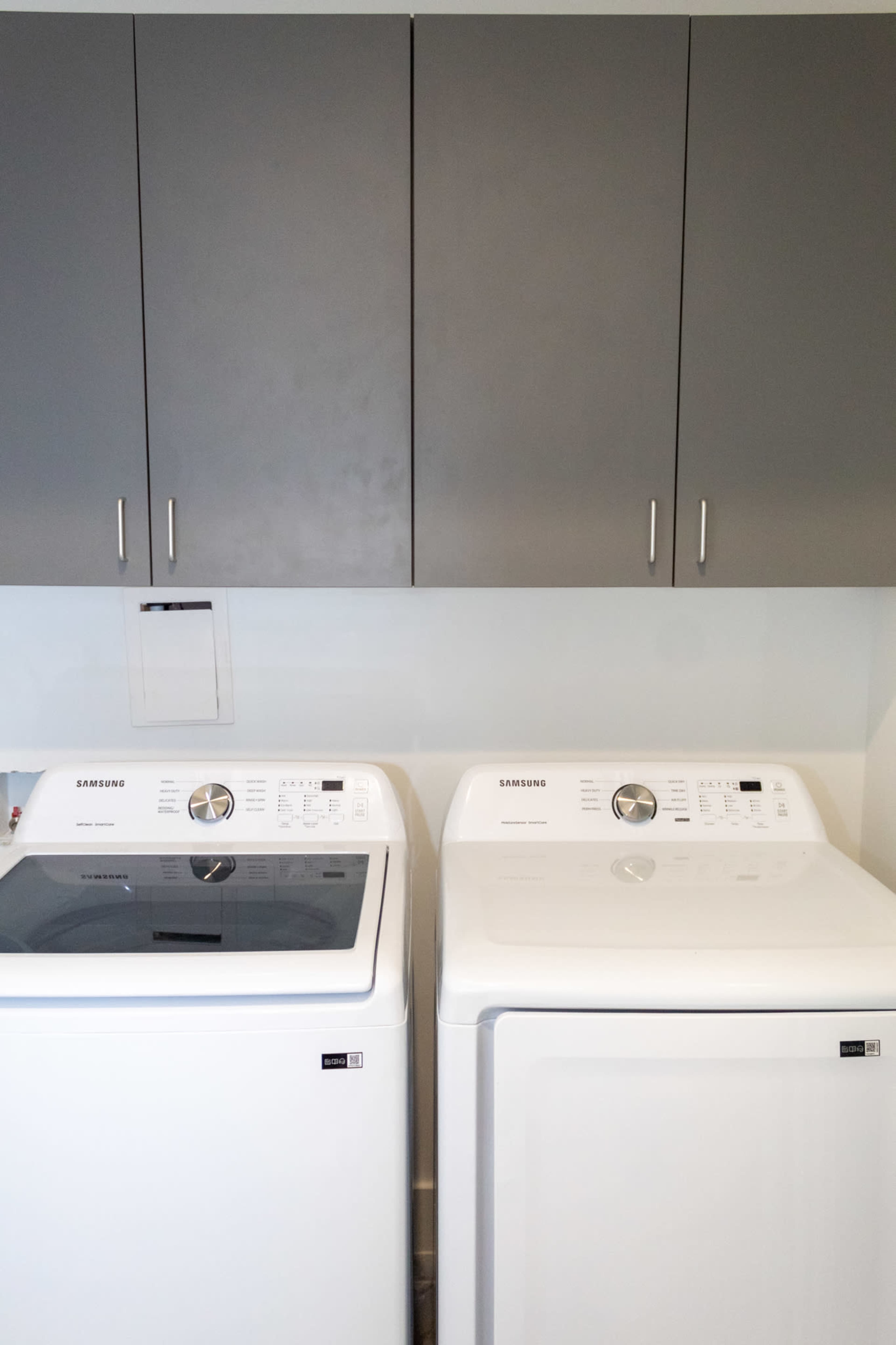 The image shows a side-by-side arrangement of a top-loading washer and a front-loading dryer beneath dark gray cabinets.