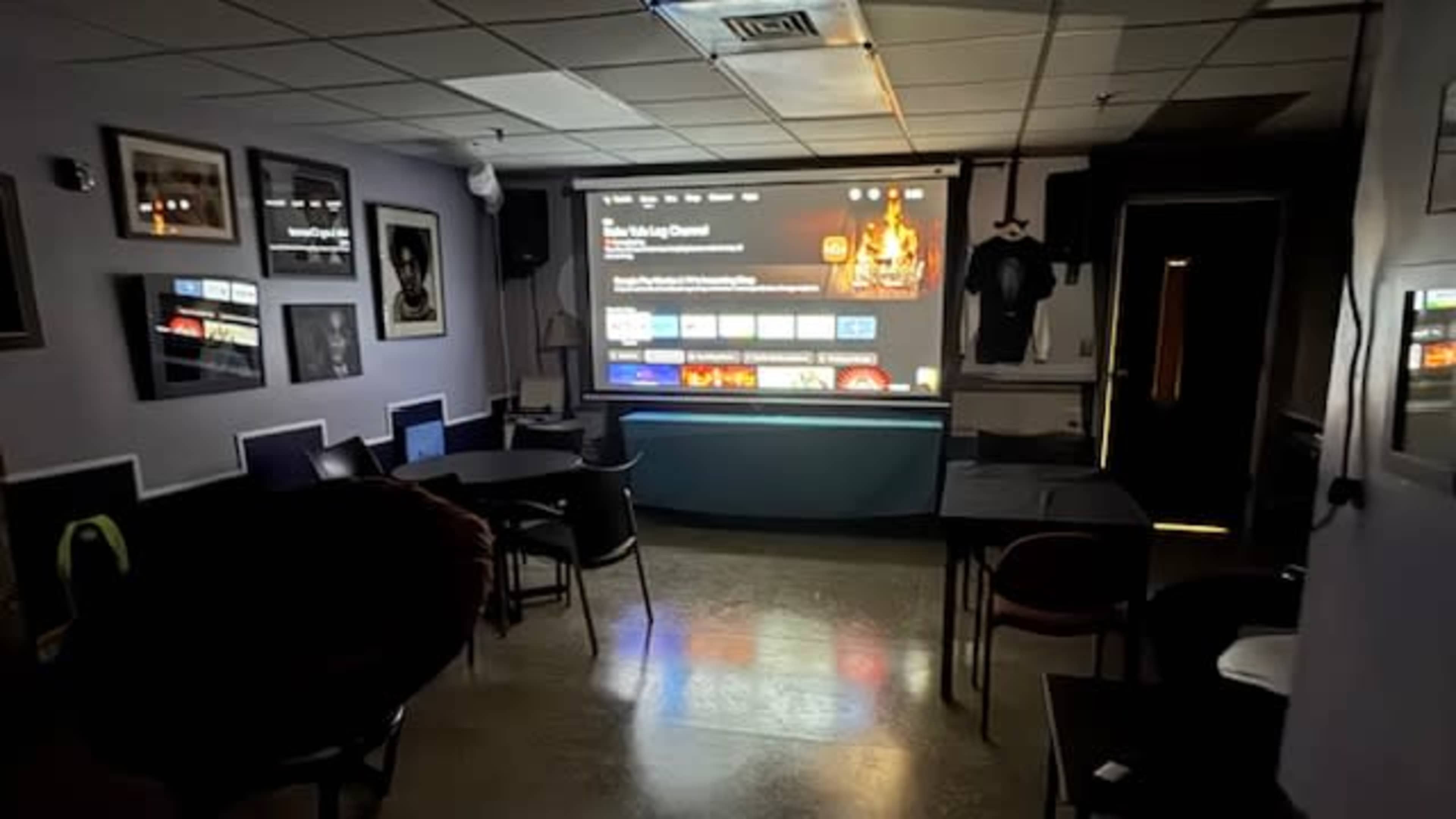 A dimly lit room features a large projection screen displaying streaming content, surrounded by various framed pictures and seating areas.