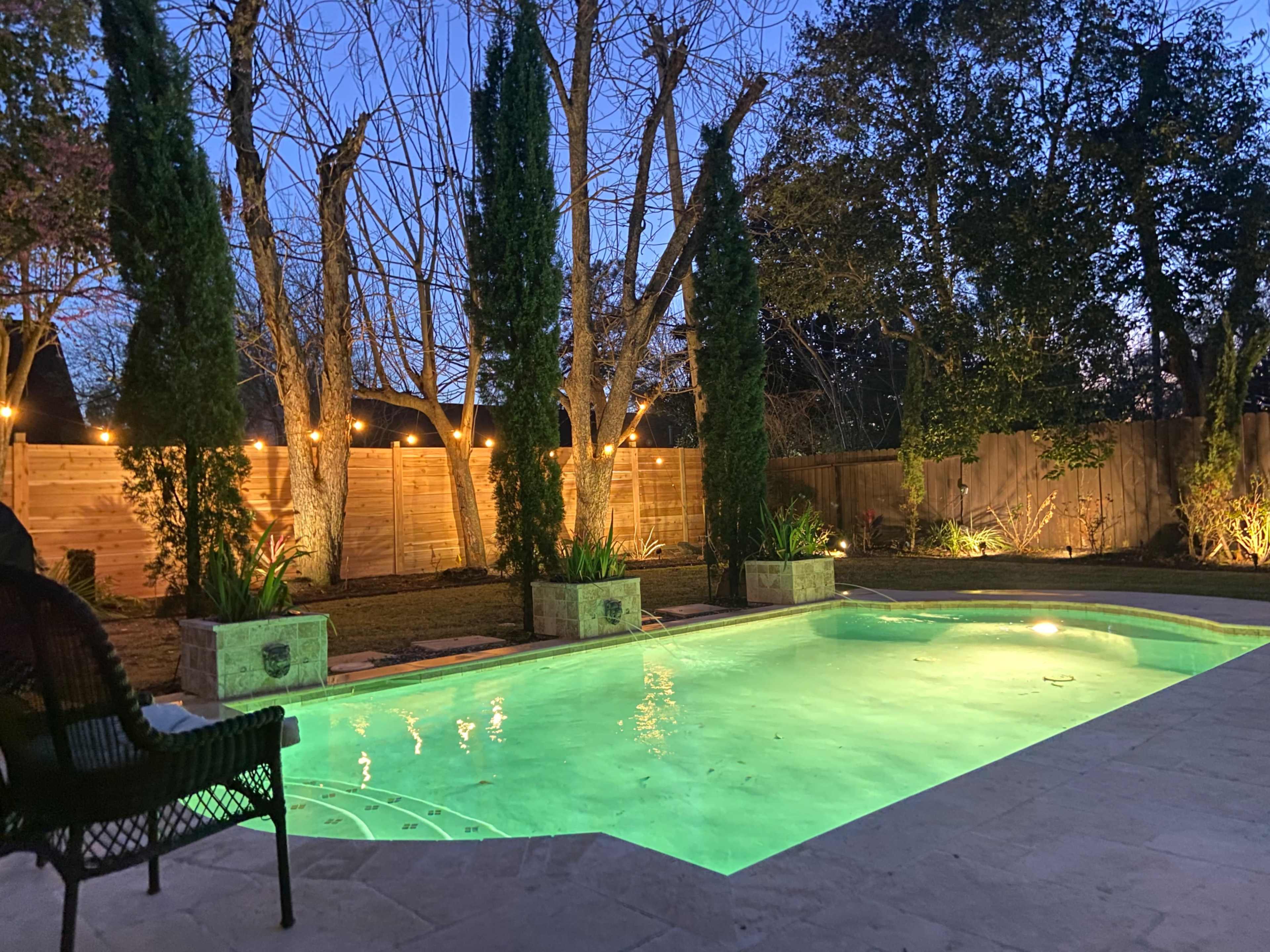 The scene shows a backyard pool surrounded by tall trees and plants, illuminated by landscape lighting at dusk.