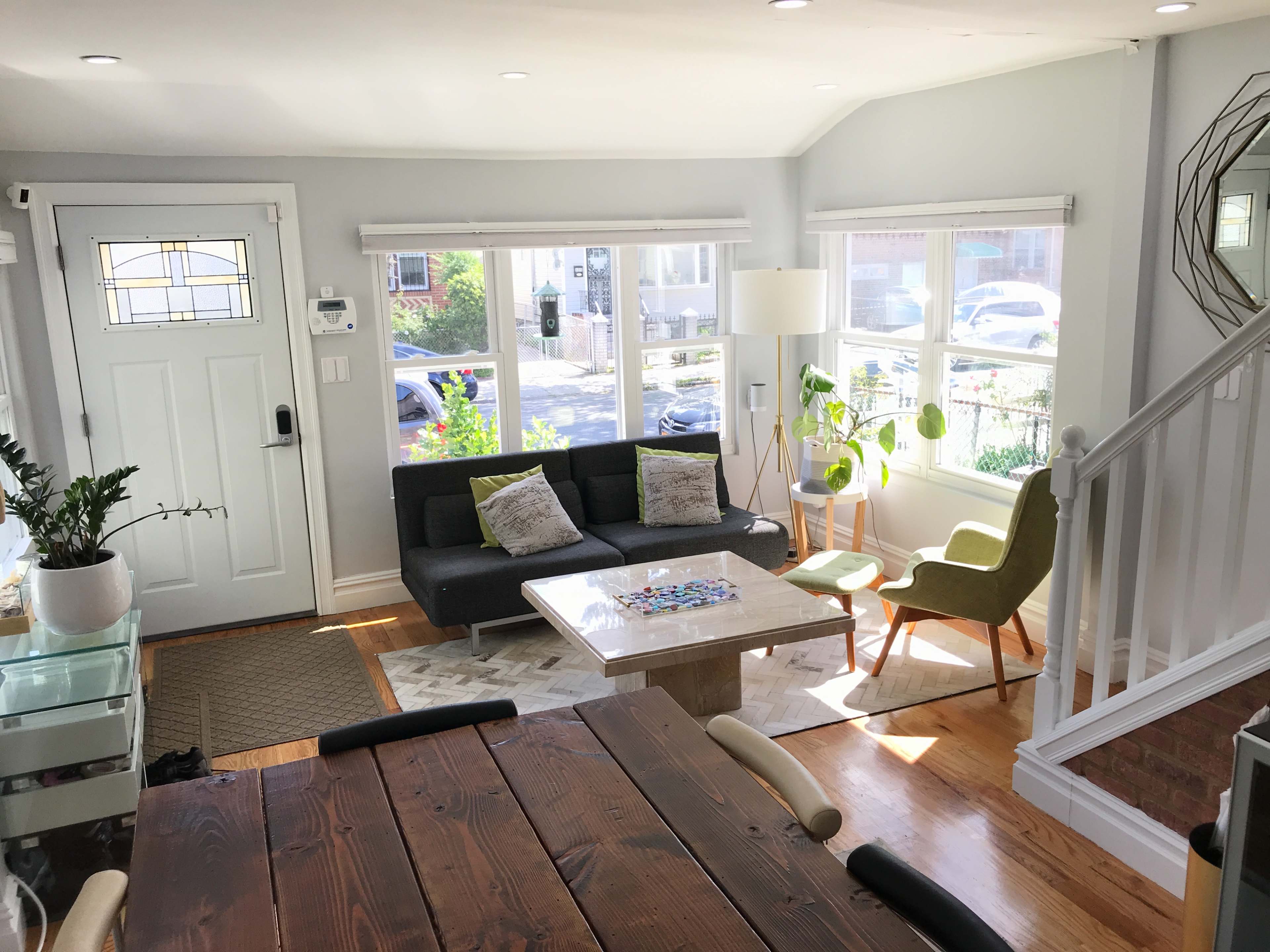 The image depicts a light-filled living room with a sofa and a coffee table, featuring a view through large windows and a staircase in the background.