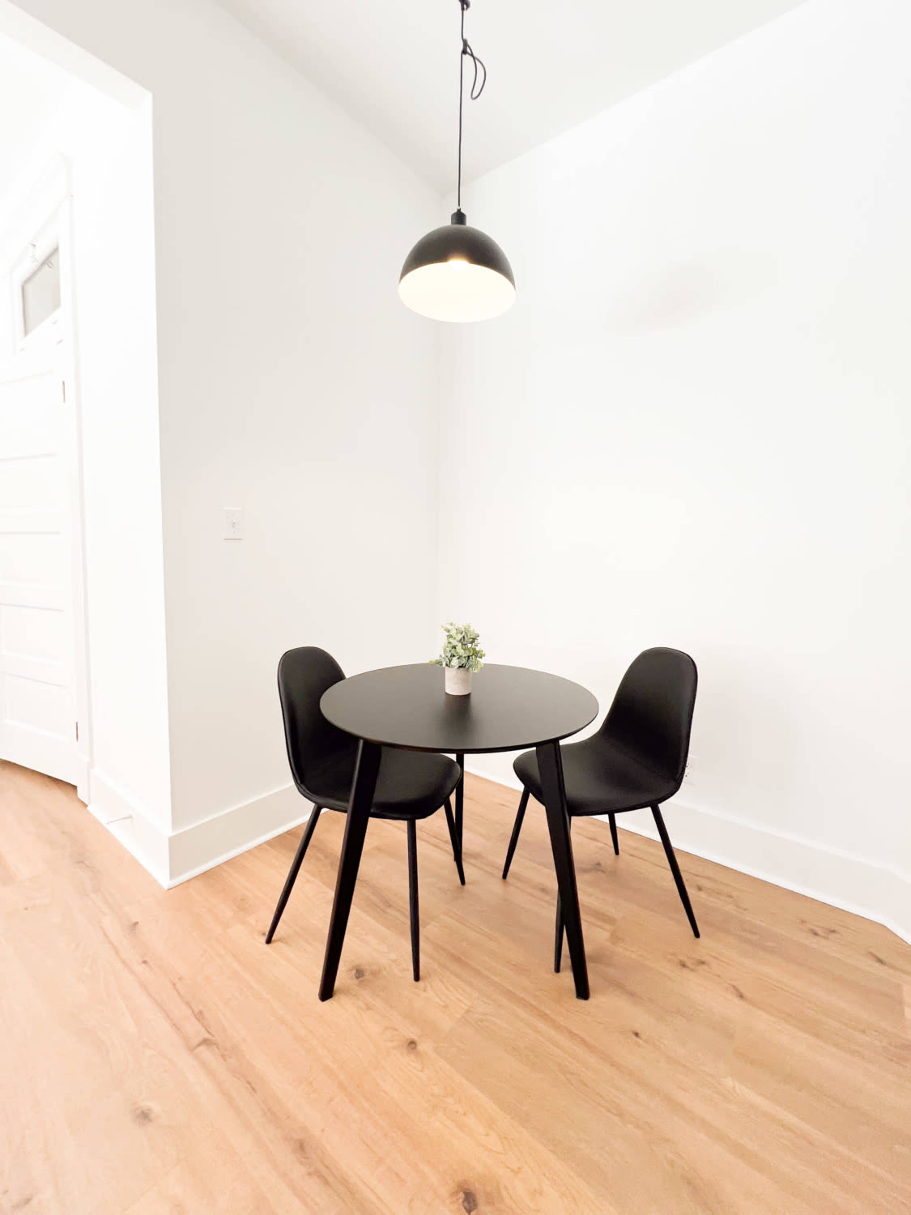 A small round table with two black chairs is positioned on a wooden floor, accompanied by a potted plant on the table under a hanging light fixture.