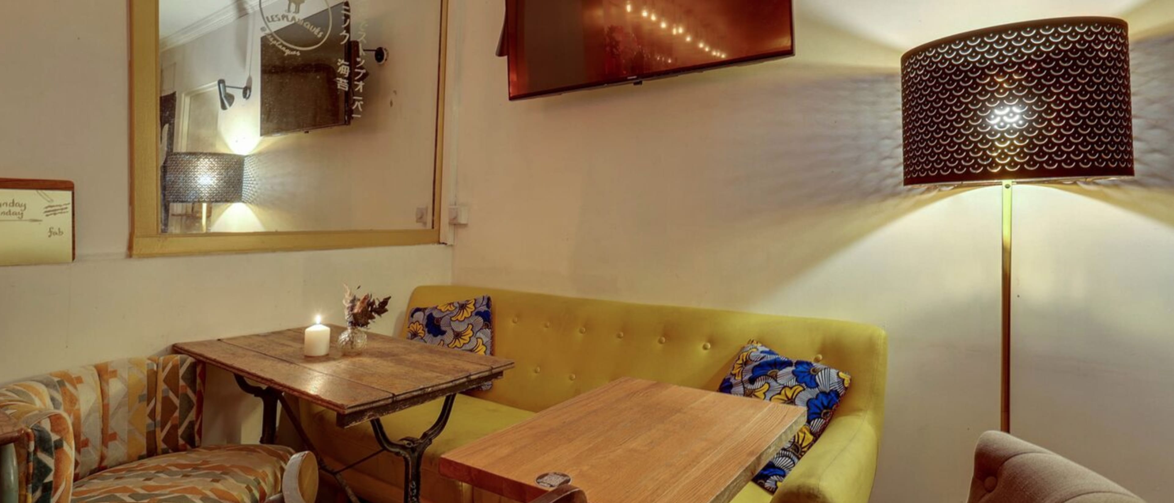 A cozy seating area with a yellow sofa, a wooden table, and a patterned armchair, illuminated by a lamp and a television mounted on the wall.