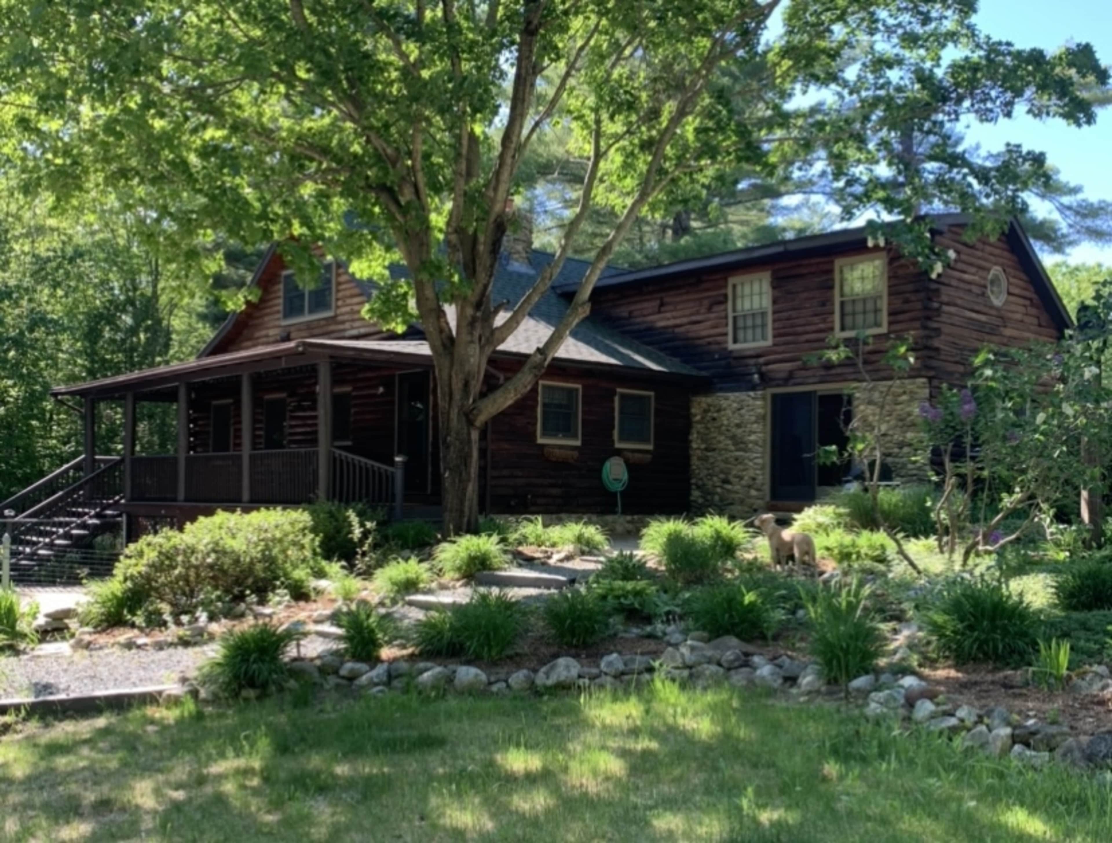 A wooden house with a stone section and a porch is surrounded by greenery and trees.