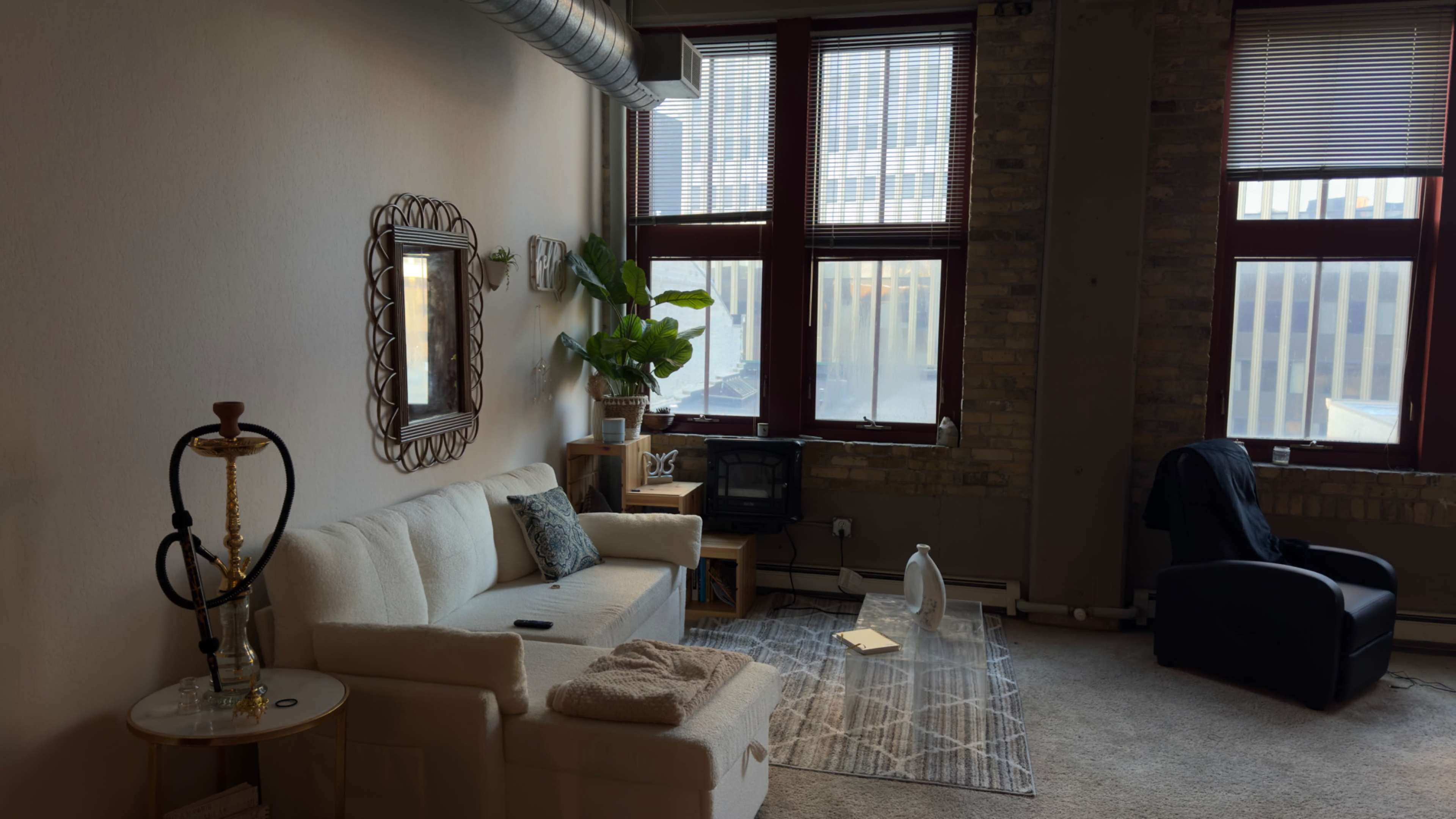 A cozy living room with a light-colored sofa, a potted plant, a small table, and large windows overlooking a cityscape.
