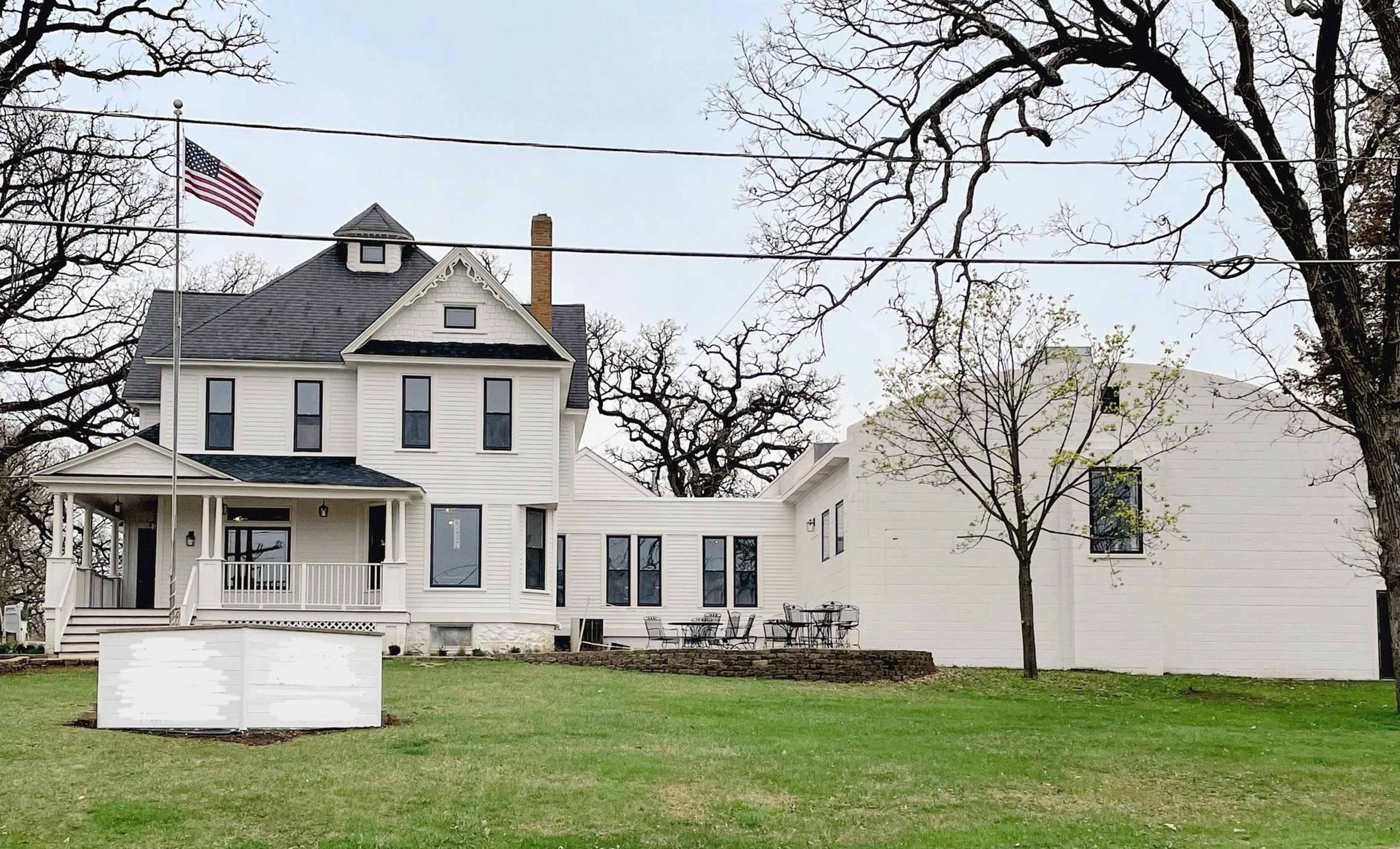 A large two-story house with a flagpole stands next to a smaller one-story building on a grassy lawn.