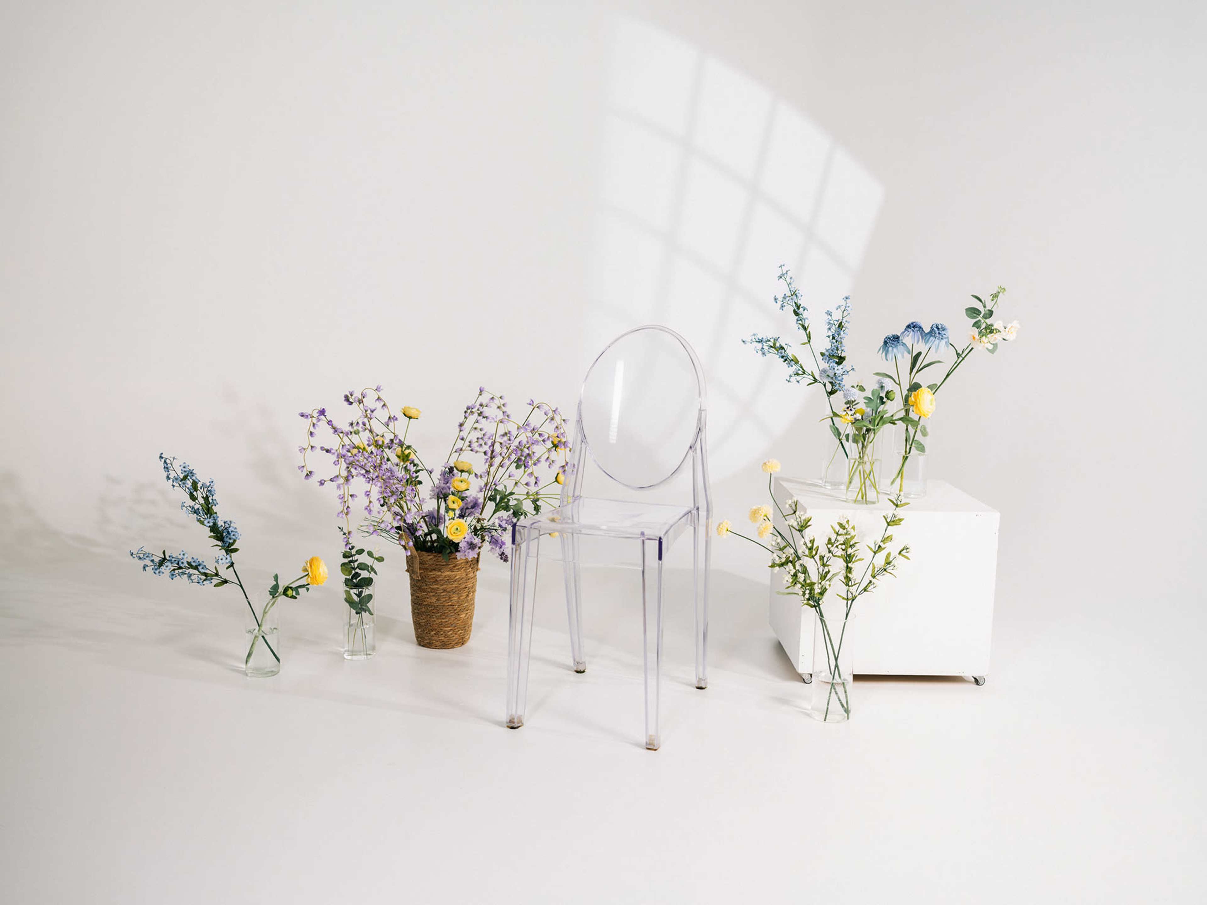 The image features a transparent chair surrounded by various vases of flowers arranged on a light-colored backdrop.