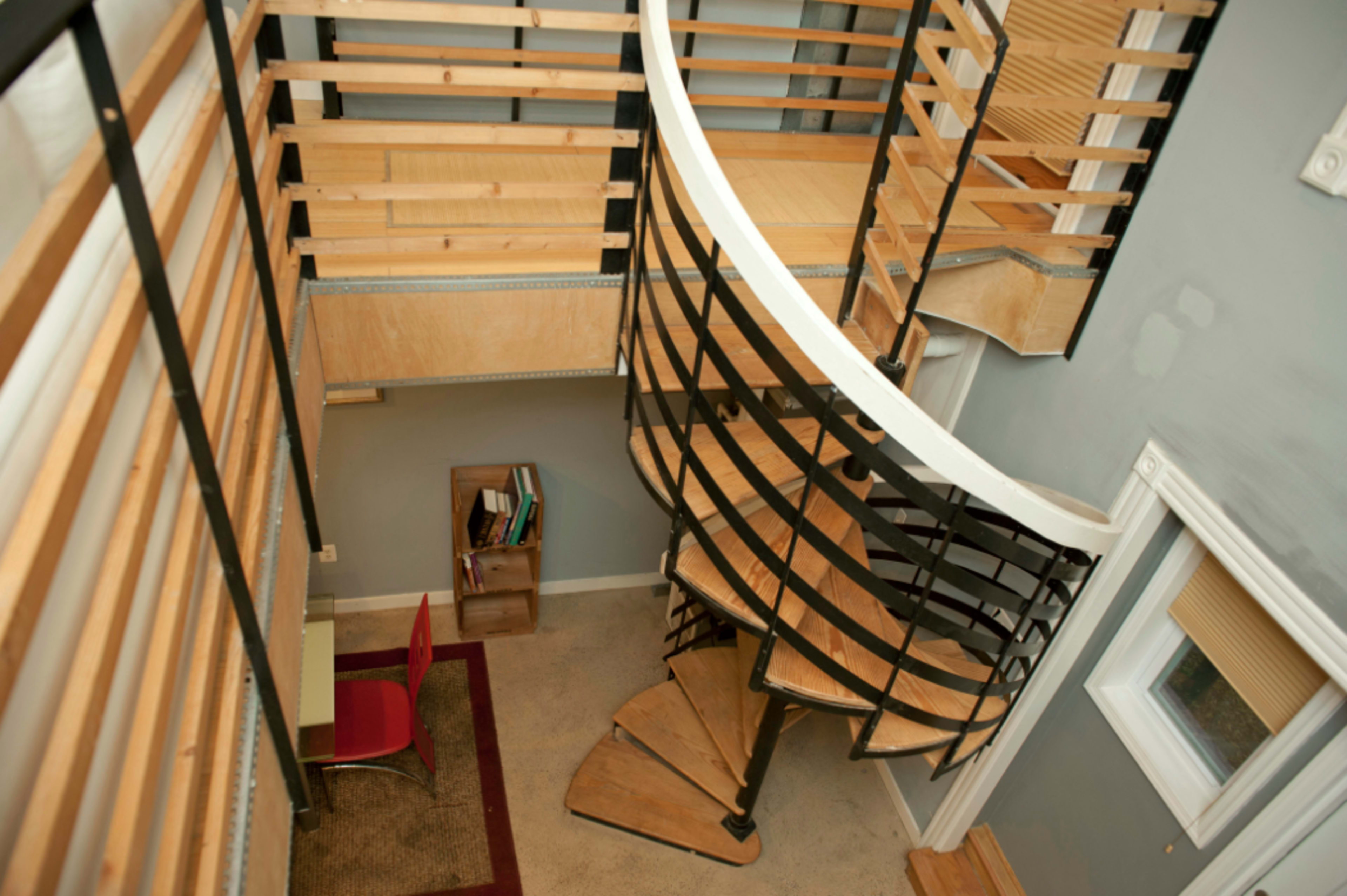 The image shows a two-story interior space featuring a curved staircase, wooden railings, and a desk area with a red chair.