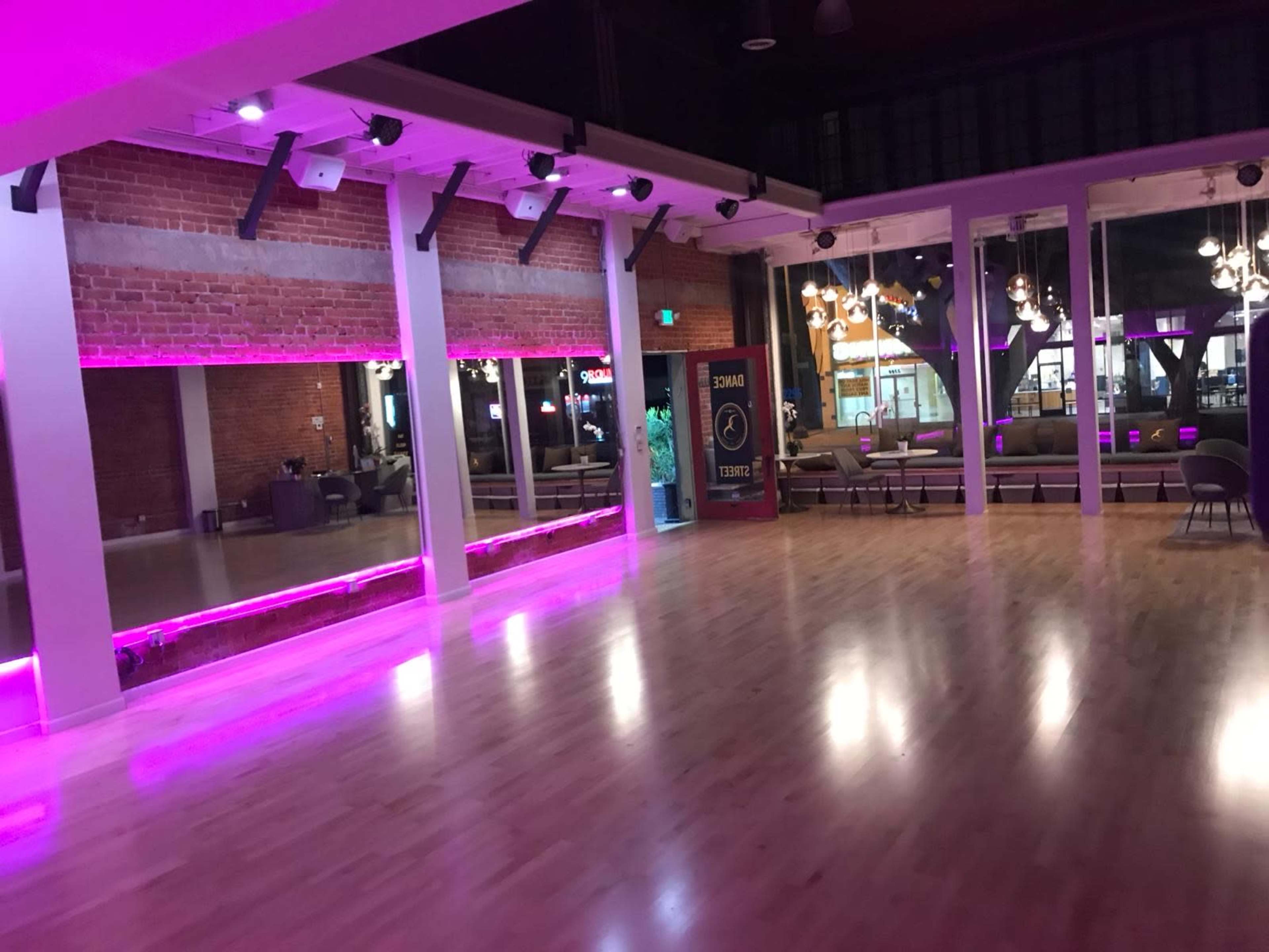 The image shows a spacious dance studio with polished wooden floors, accented by pink LED lighting along the walls and a large glass window revealing a lounge area outside.