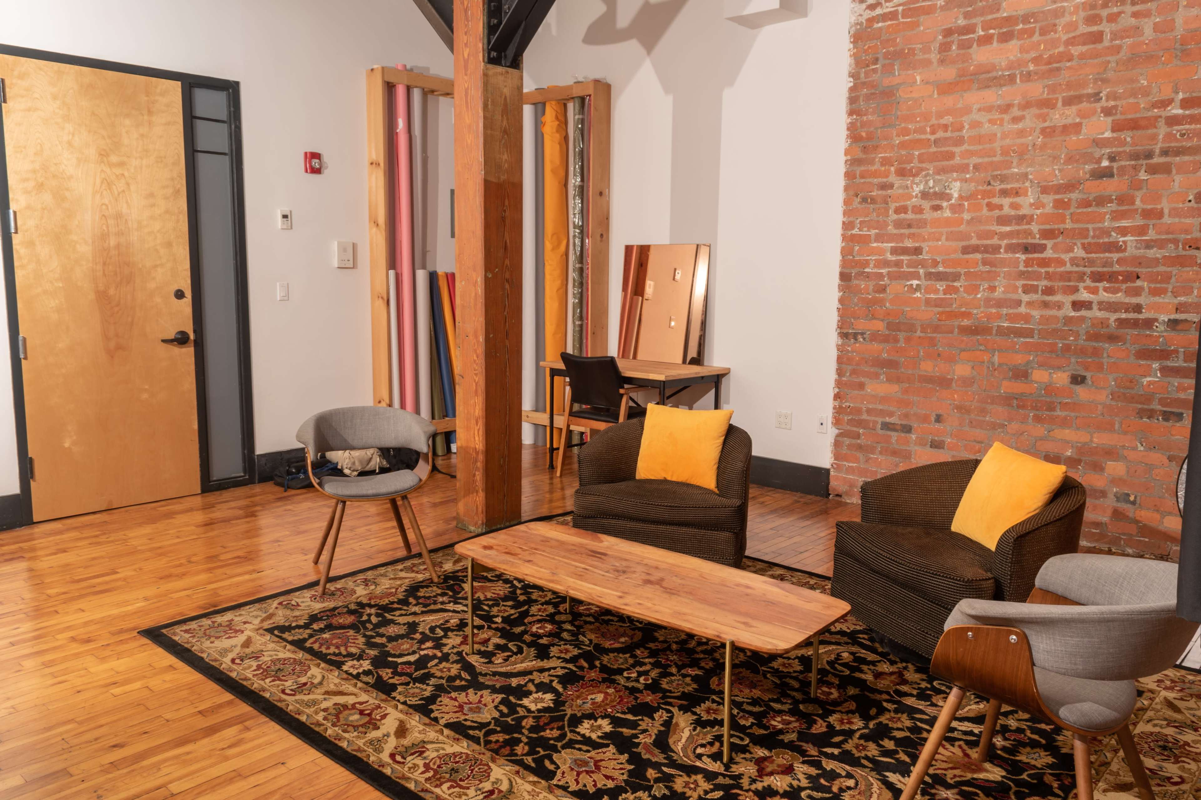 A modern interior space with a wooden floor, featuring a low coffee table, several armchairs with cushions, a mirror, and a table against a brick wall.