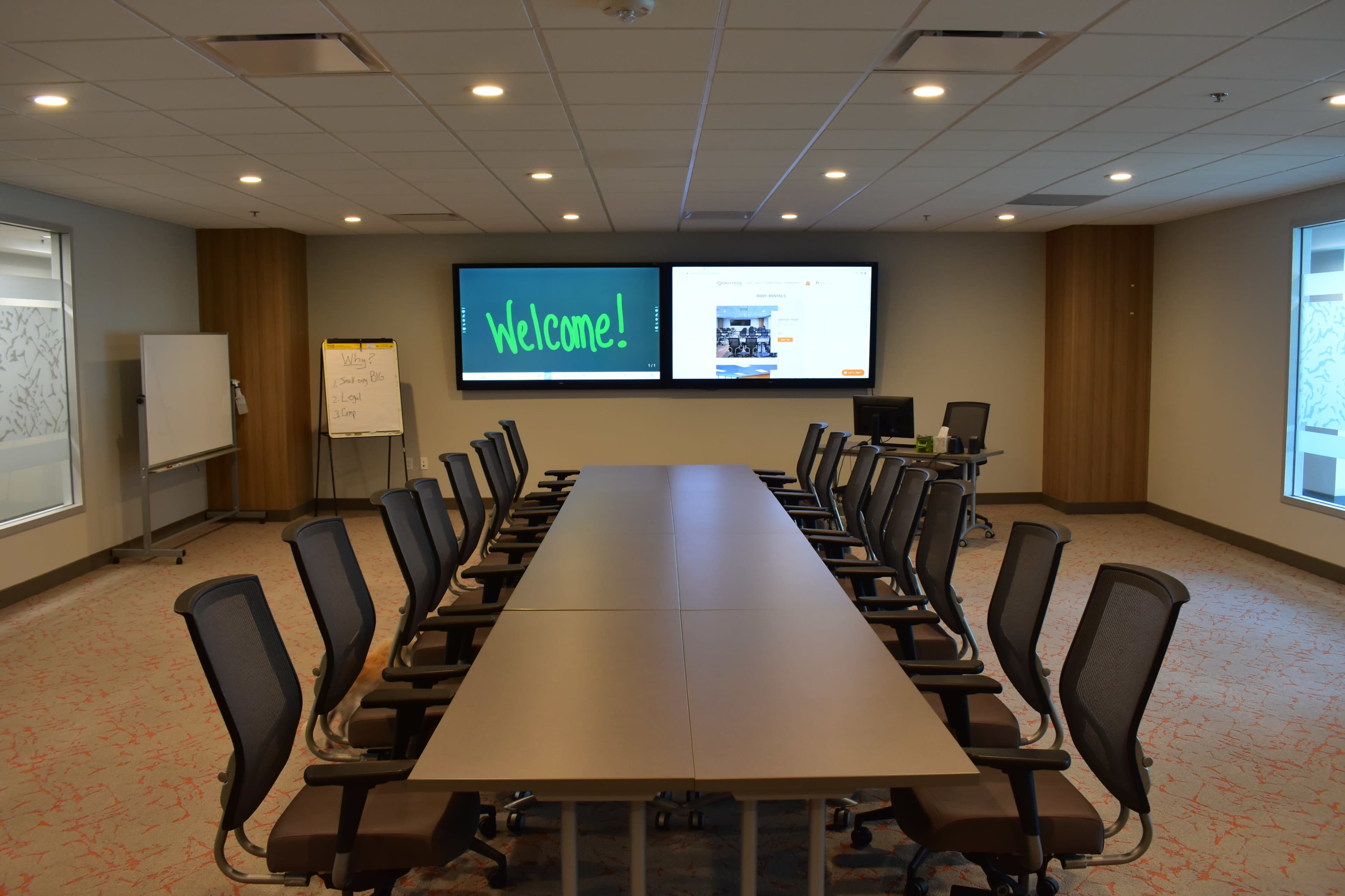 A large conference room features a long table surrounded by ergonomic chairs, two screens displaying a welcoming message and a presentation, and a whiteboard in the corner.