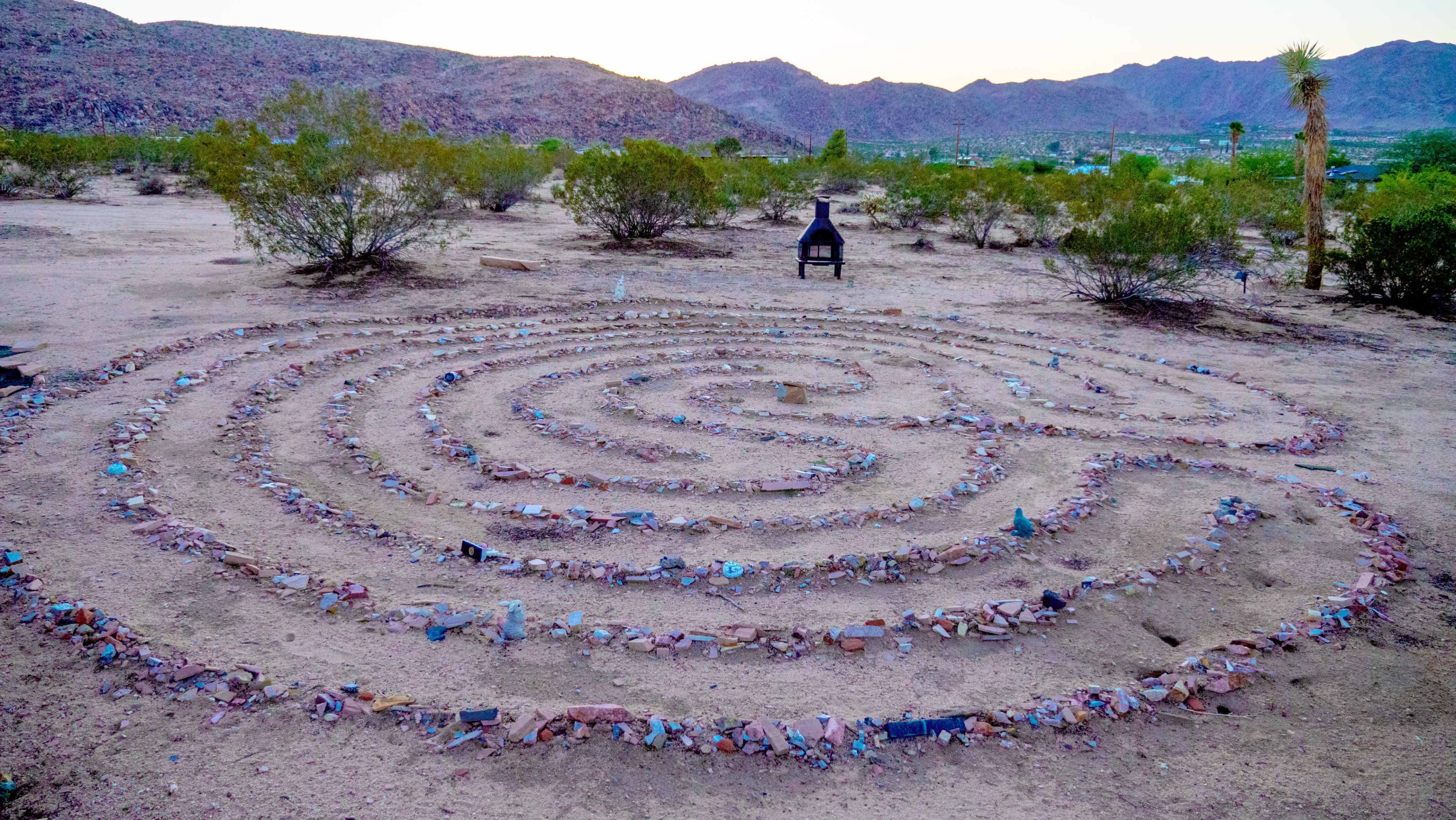 A circular stone labyrinth is situated in a desert landscape, surrounded by sparse shrubs and mountains in the background.