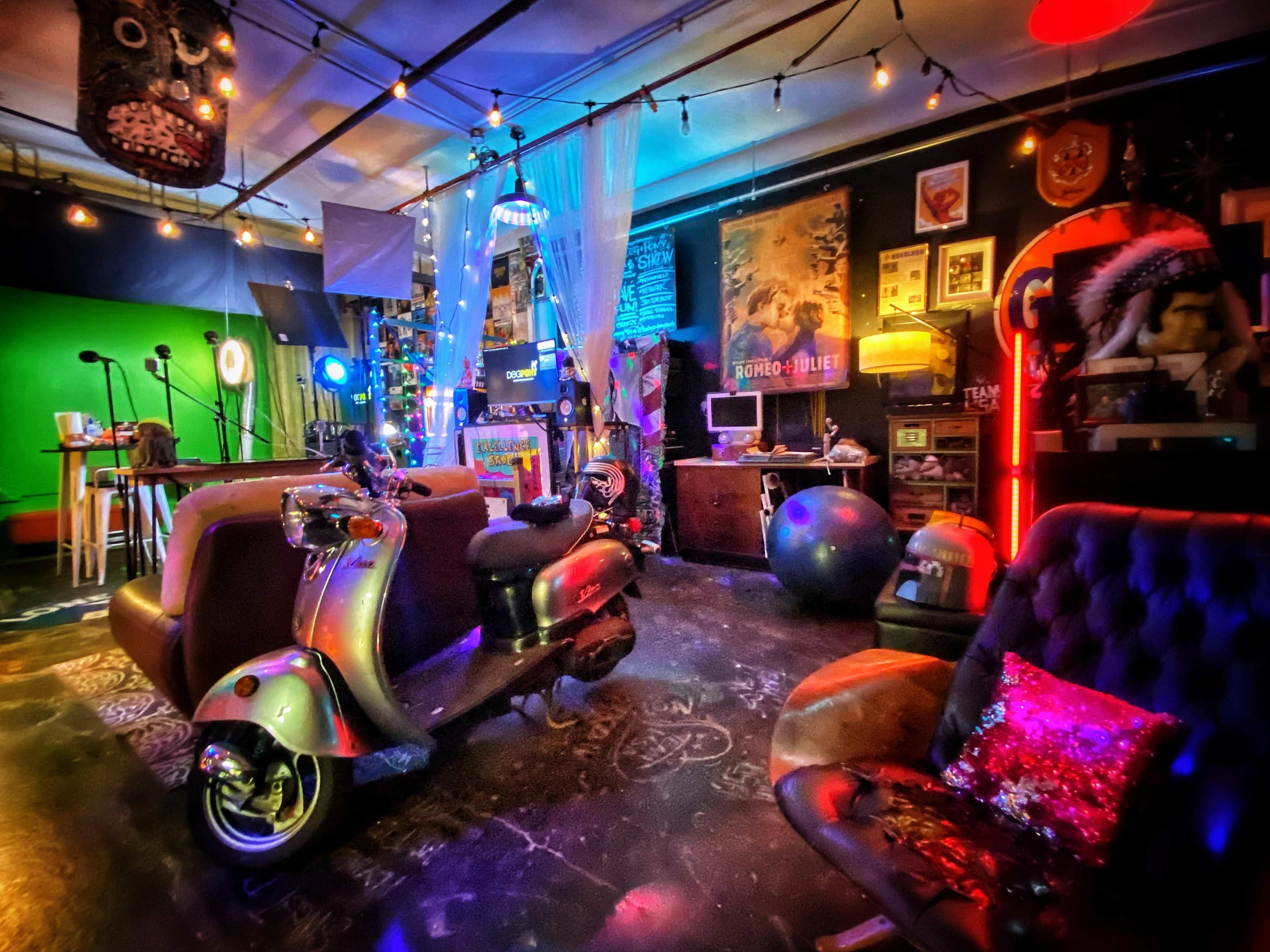 The scene features a colorful room with retro furniture, a scooter, and various art and lighting elements, including a green screen and string lights.