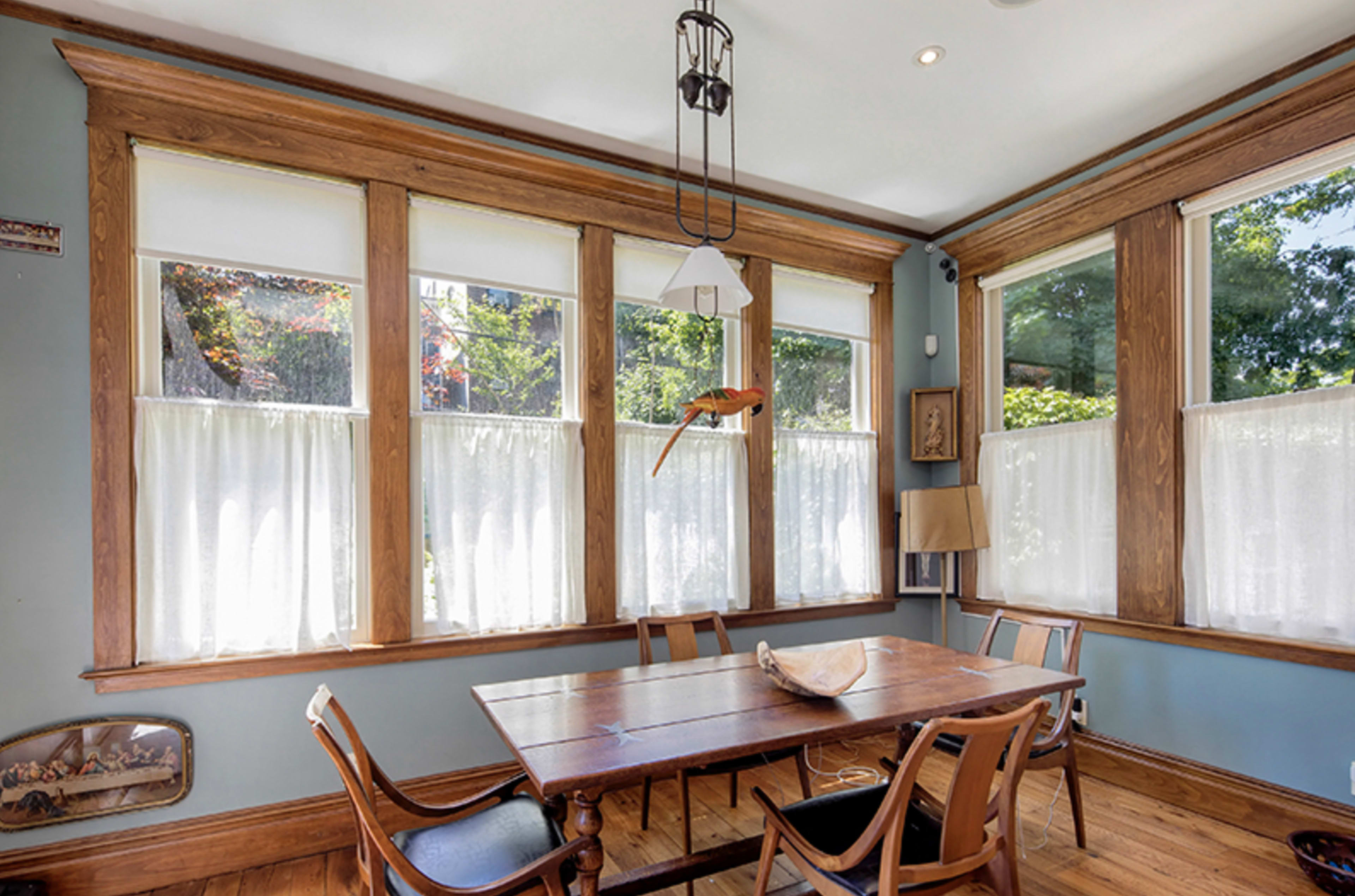 A dining area features a wooden table surrounded by chairs, with large windows dressed in sheer curtains allowing natural light to fill the space.